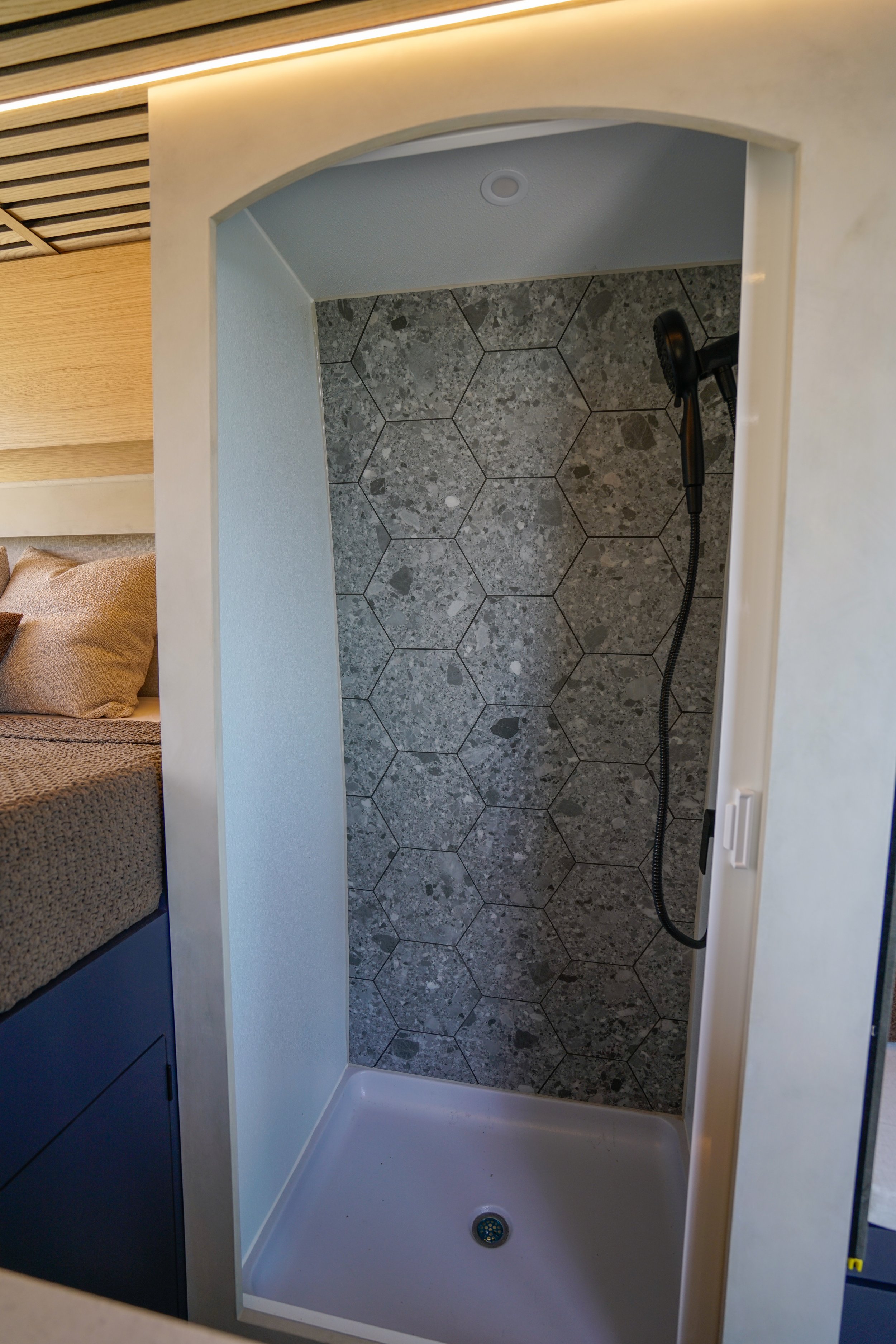 Empty walk-in shower with hexagon gray tile walls and a handheld showerhead, viewed from the entrance.