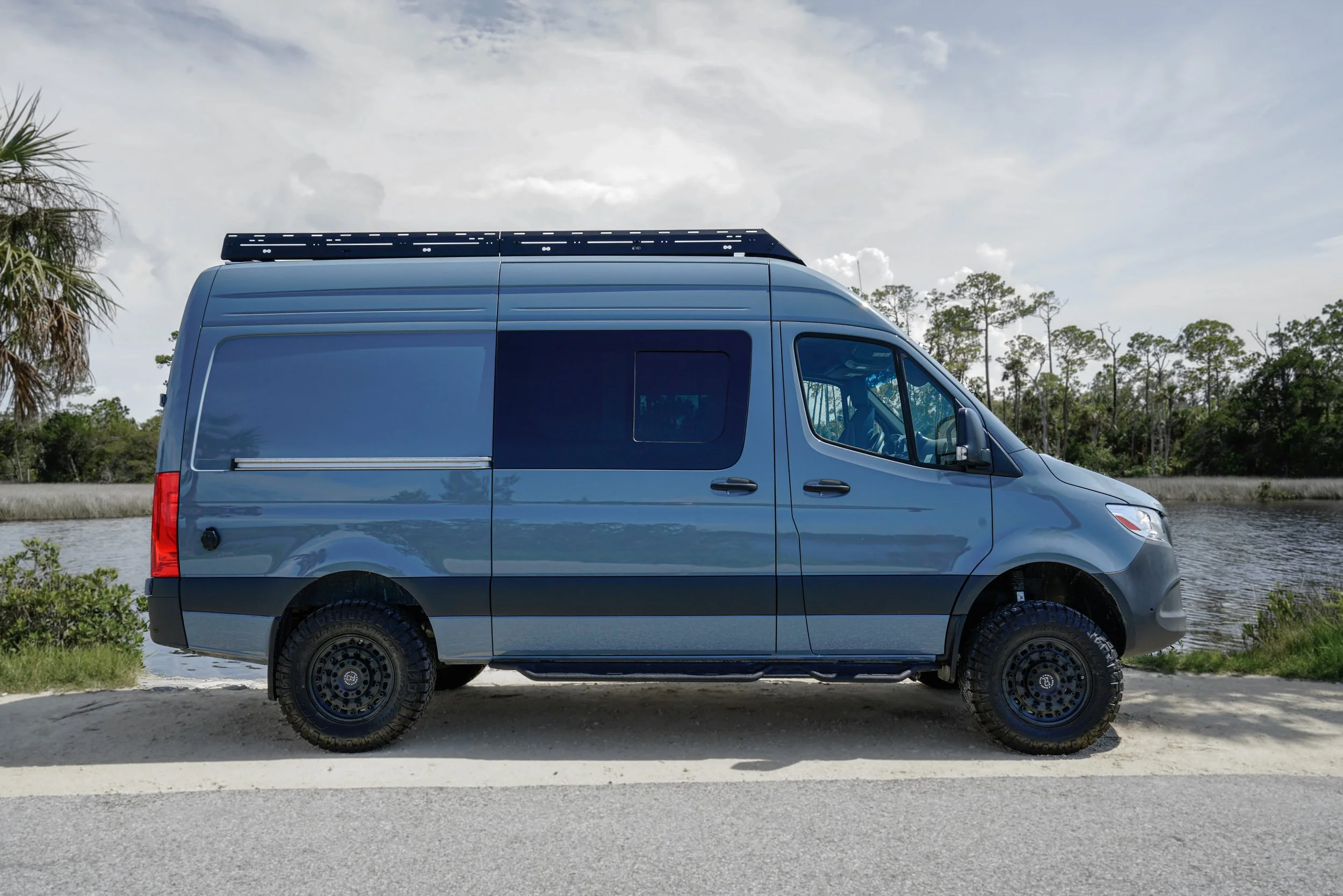 A gray sprinter van parked outdoors near water with trees and cloudy sky in the background.