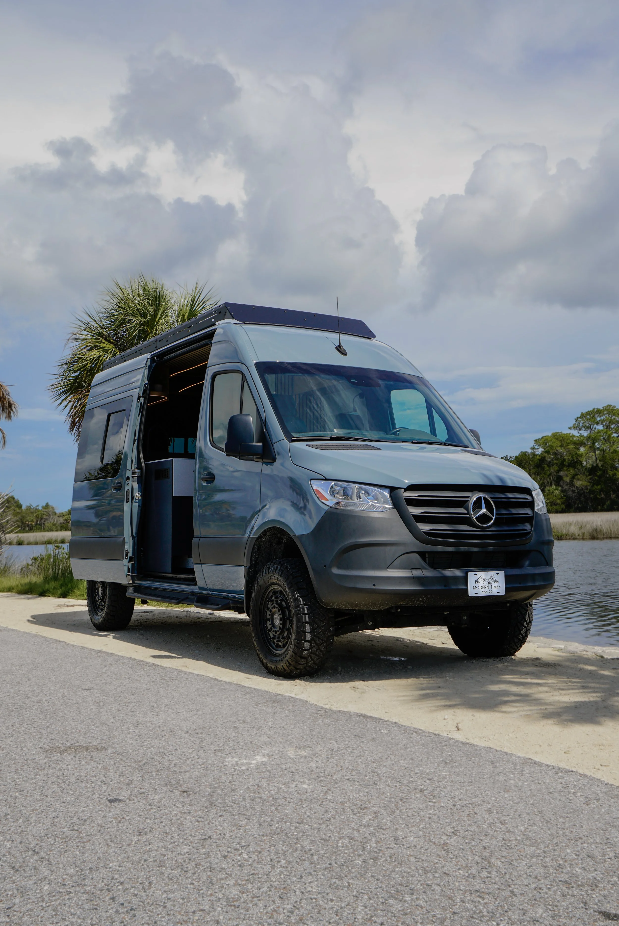 A grey Mercedes-Benz camper van parked on a paved area near calm water with grass and trees in the background, under a cloudy sky.
