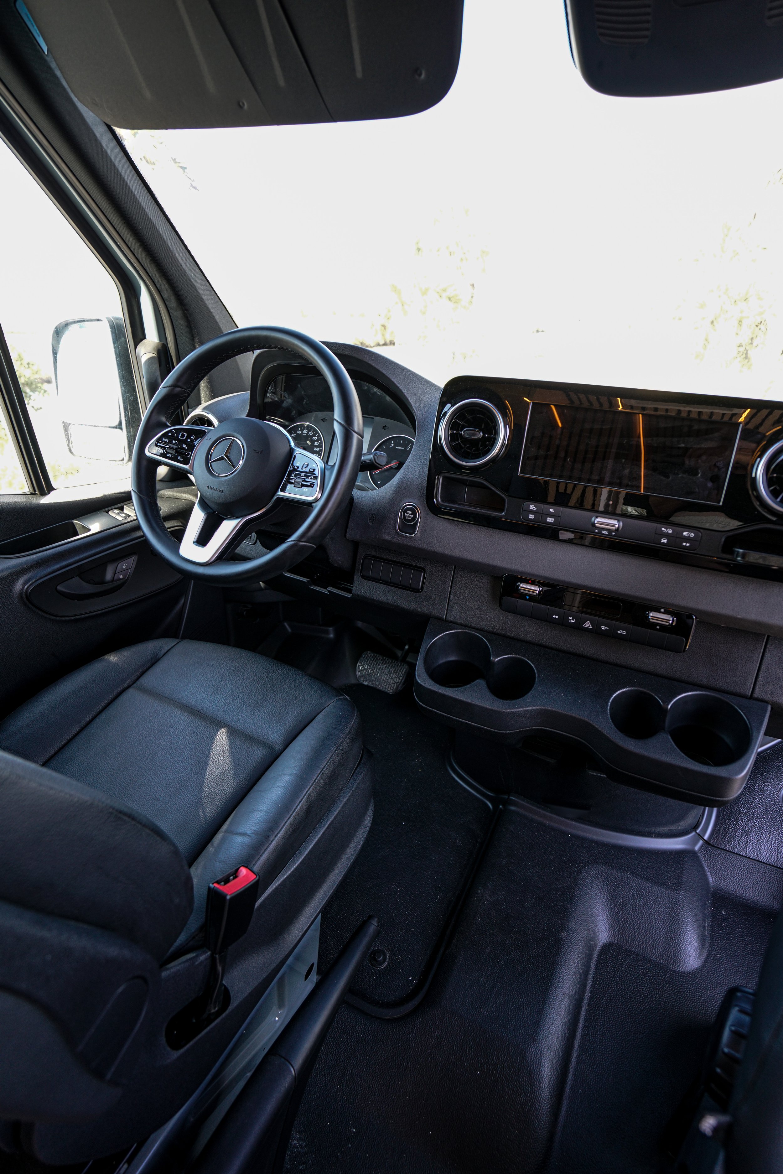 Interior view of a vehicle dashboard, steering wheel, and front seats, with a large, bright windshield showing outside scenery.