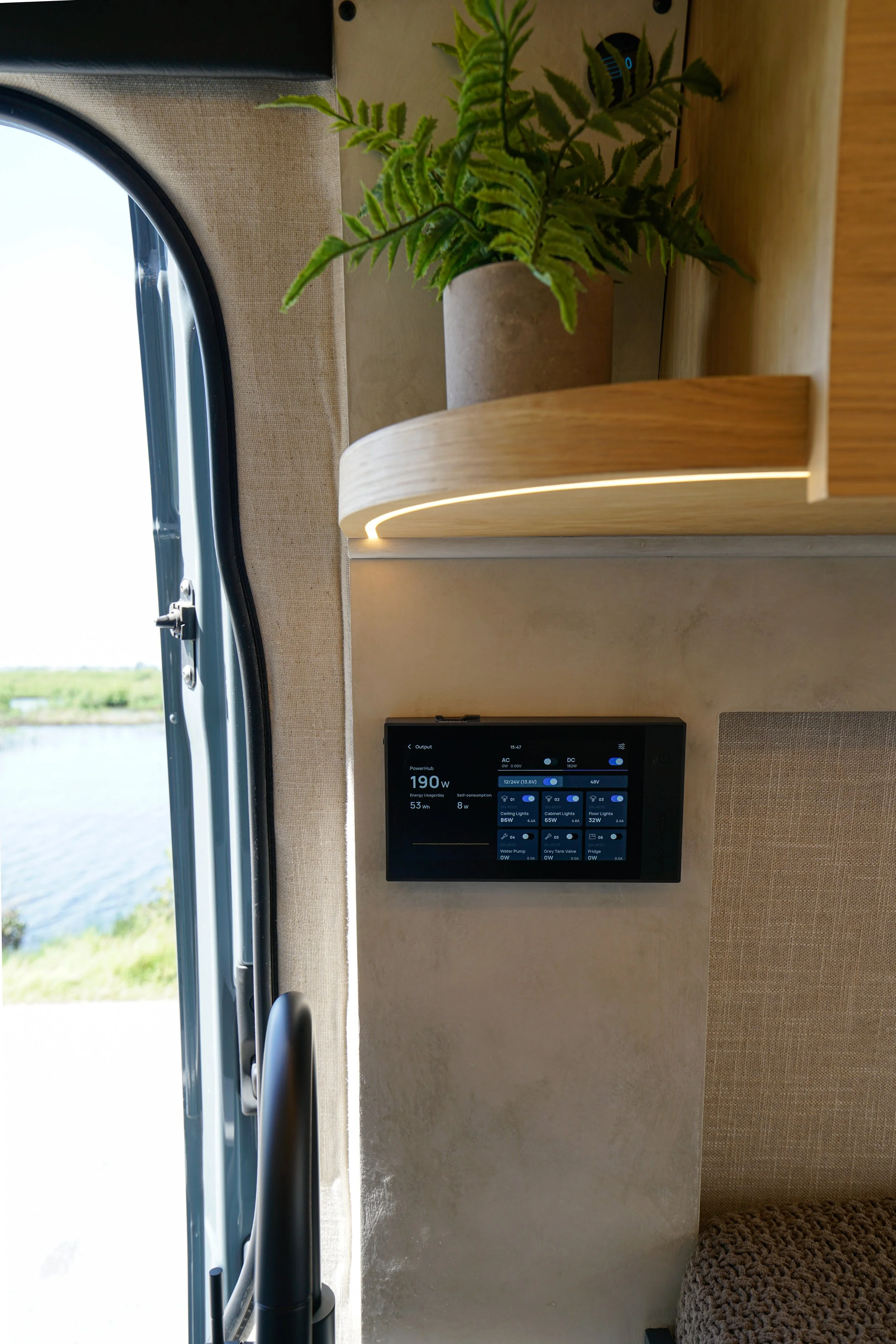 Interior of a vehicle with a potted fern on a wooden shelf and a digital control panel on the wall, with a window showing an outdoor landscape.