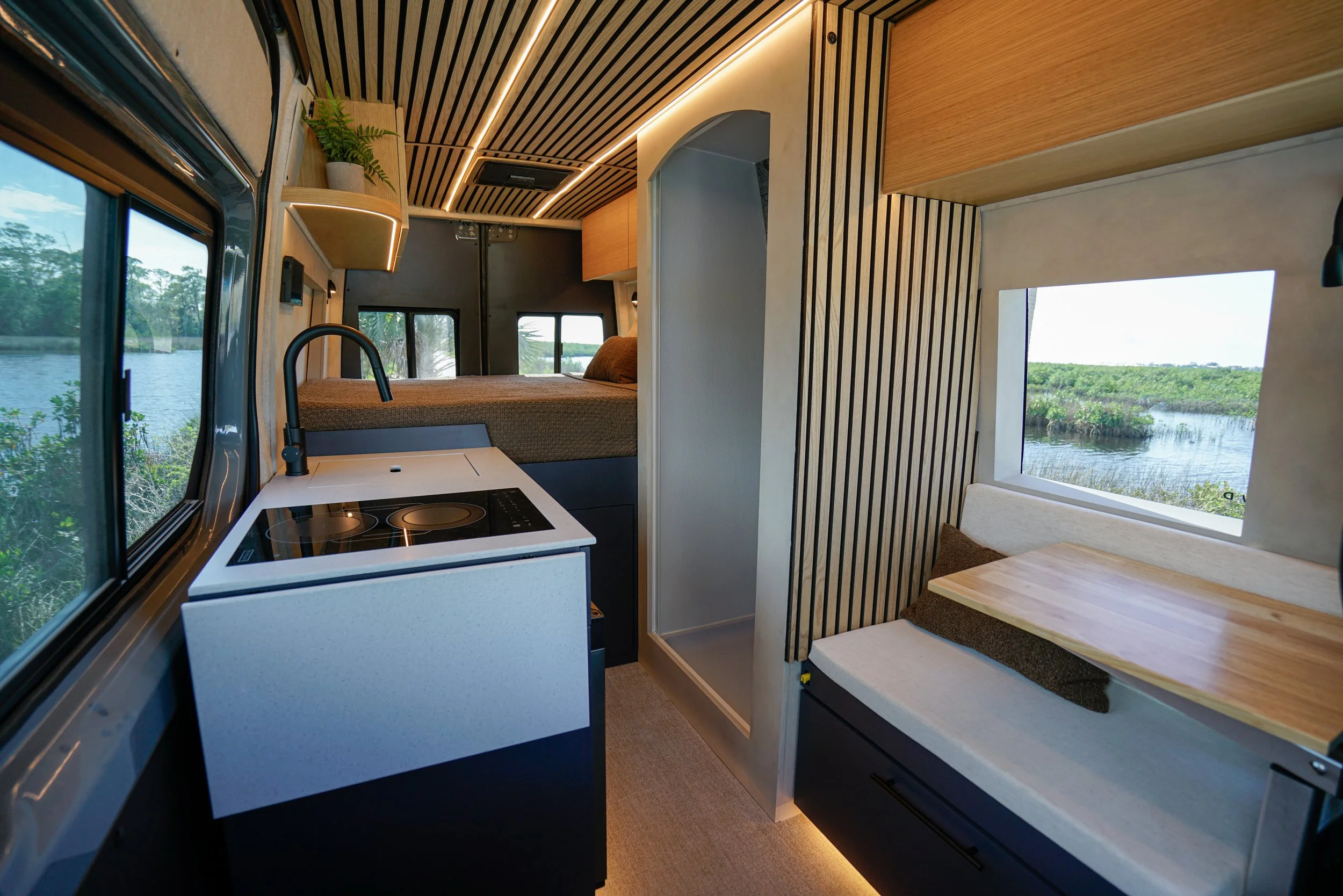 Interior of a compact camper with wooden accents, a small kitchen stove, a bed, and windows showing a green landscape and water outside.