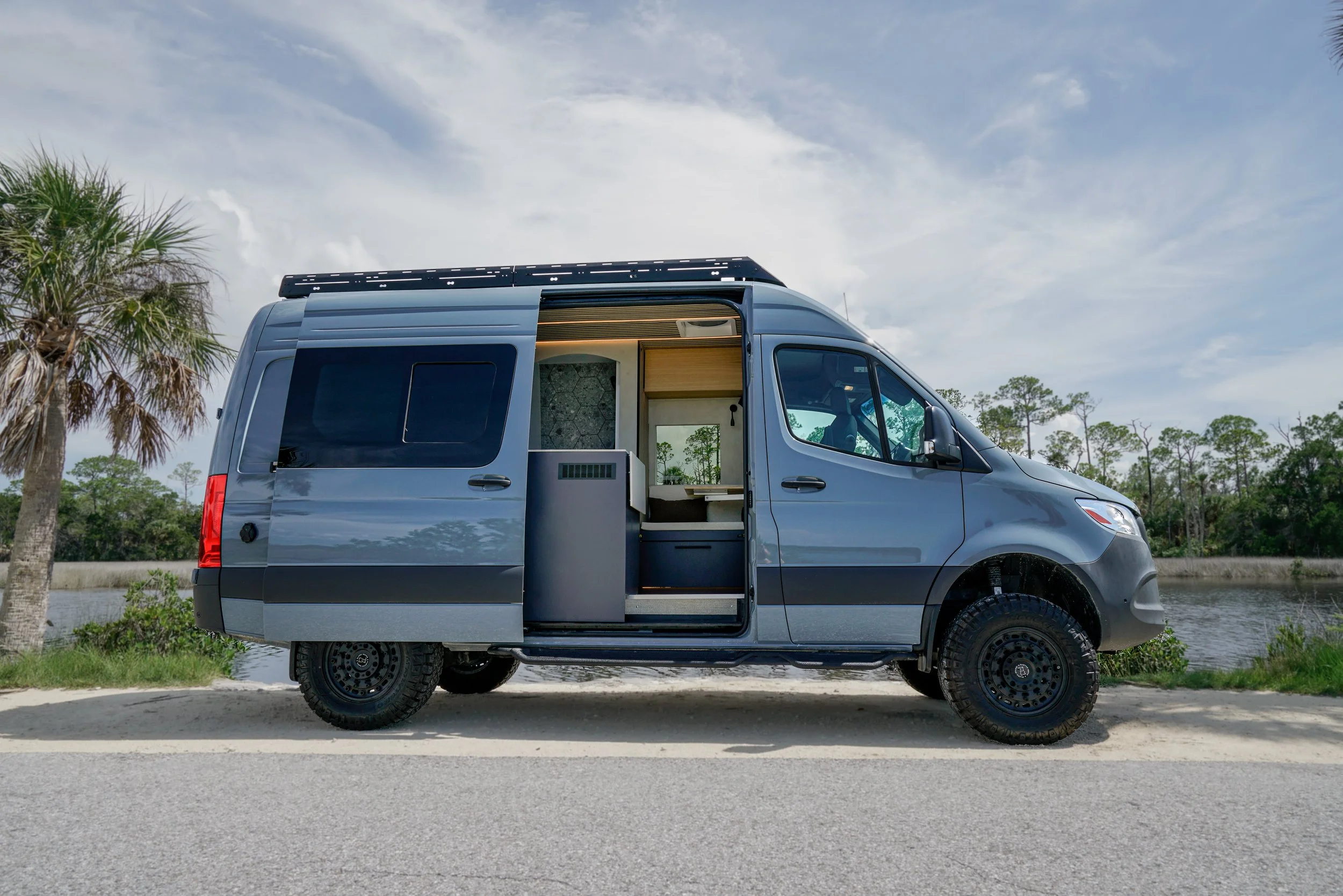 A grey camper van parked by a body of water with trees in the background and partly cloudy sky overhead.