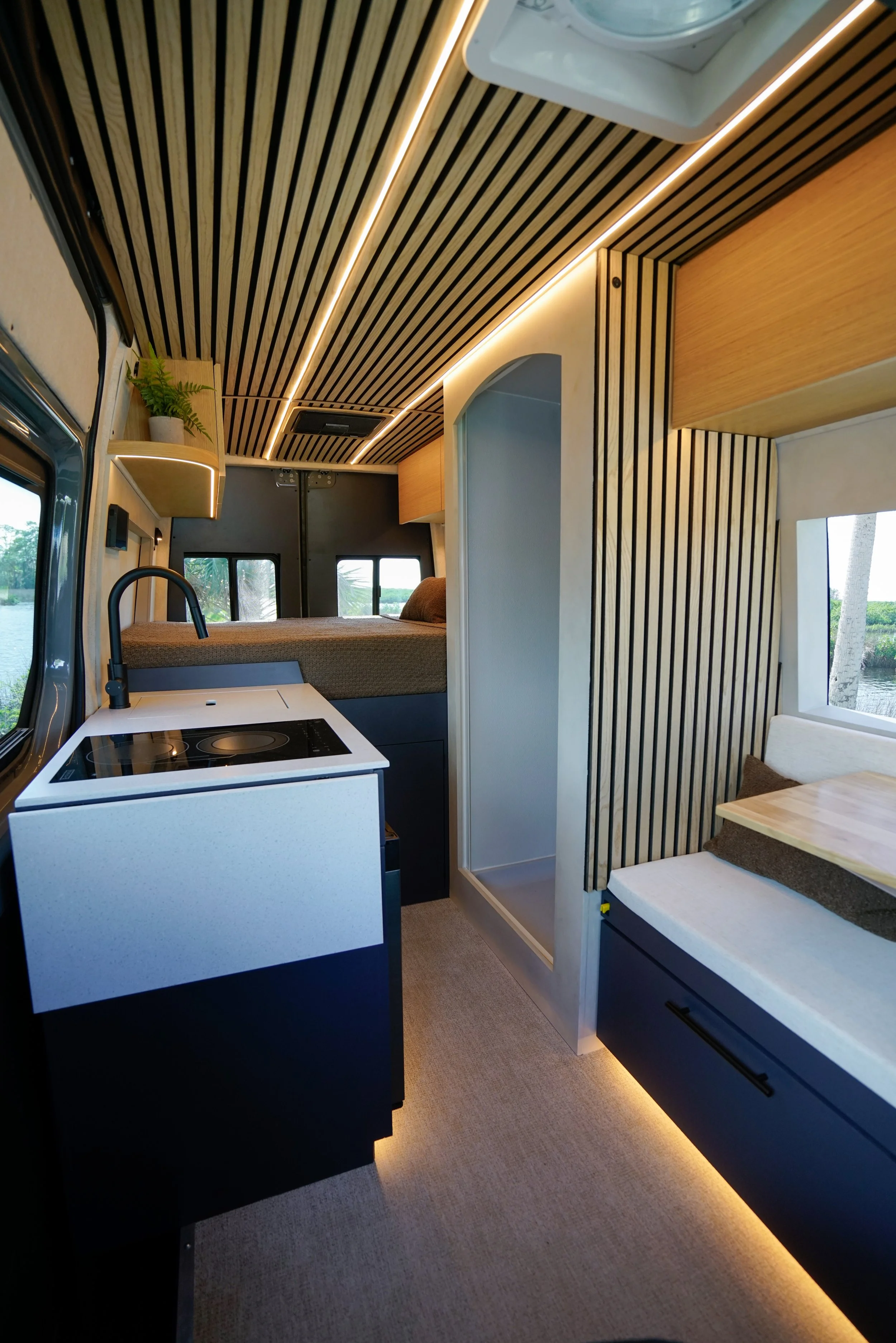 Interior of a compact living space, such as a camper van or tiny home, featuring a small kitchenette with a sink, cooktop, and minimal decor, wood paneling on the ceiling and walls, a bed in the background, and large windows showing outdoor scenery.