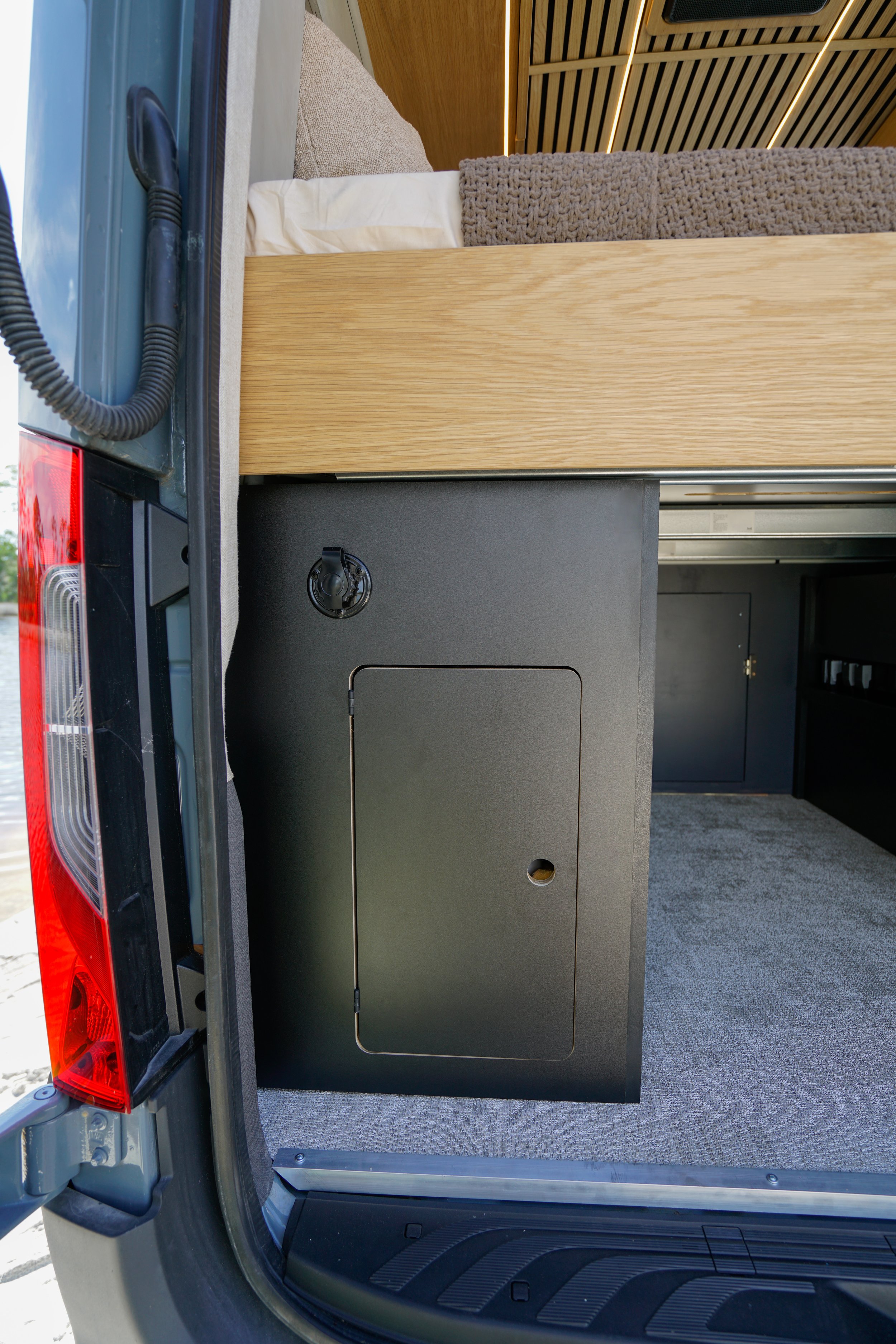 Interior of a van with a bed, gray carpet, wood paneling, black cabinetry, and a small closet with a round handle.
