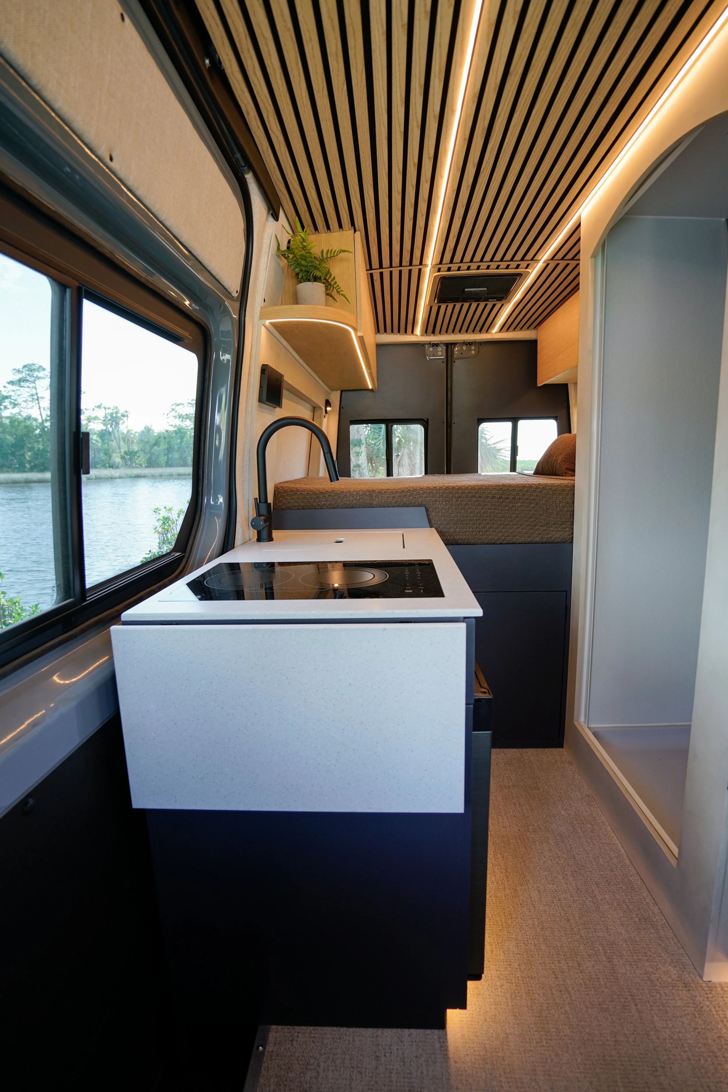 Interior of a modern tiny home or camper with a small kitchen area, bed, wooden ceiling with linear lighting, and water view through windows.