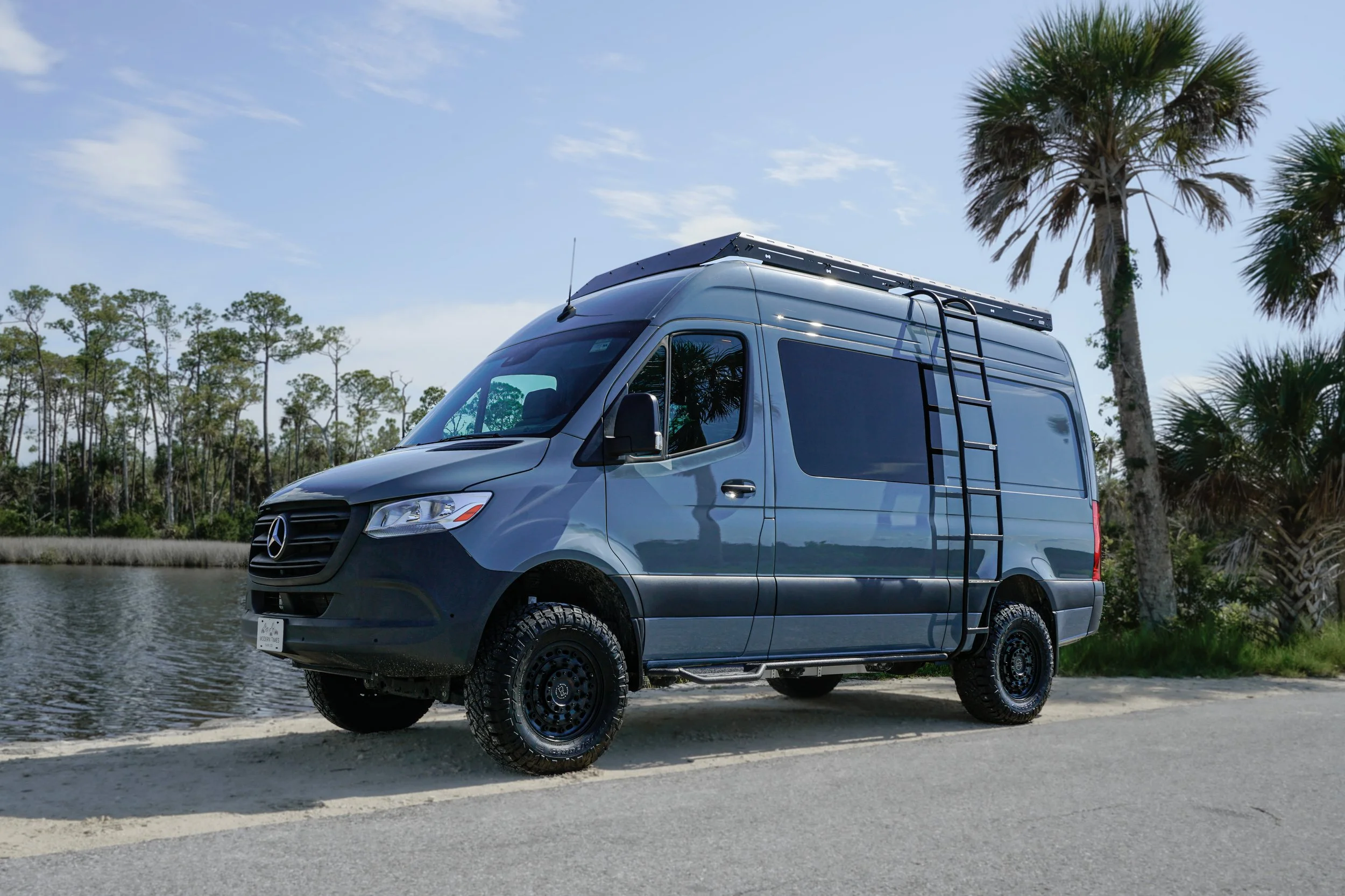A gray Mercedes-Benz Sprinter van parked on the side of a road next to a body of water with trees and palm trees in the background, under a partly cloudy sky.