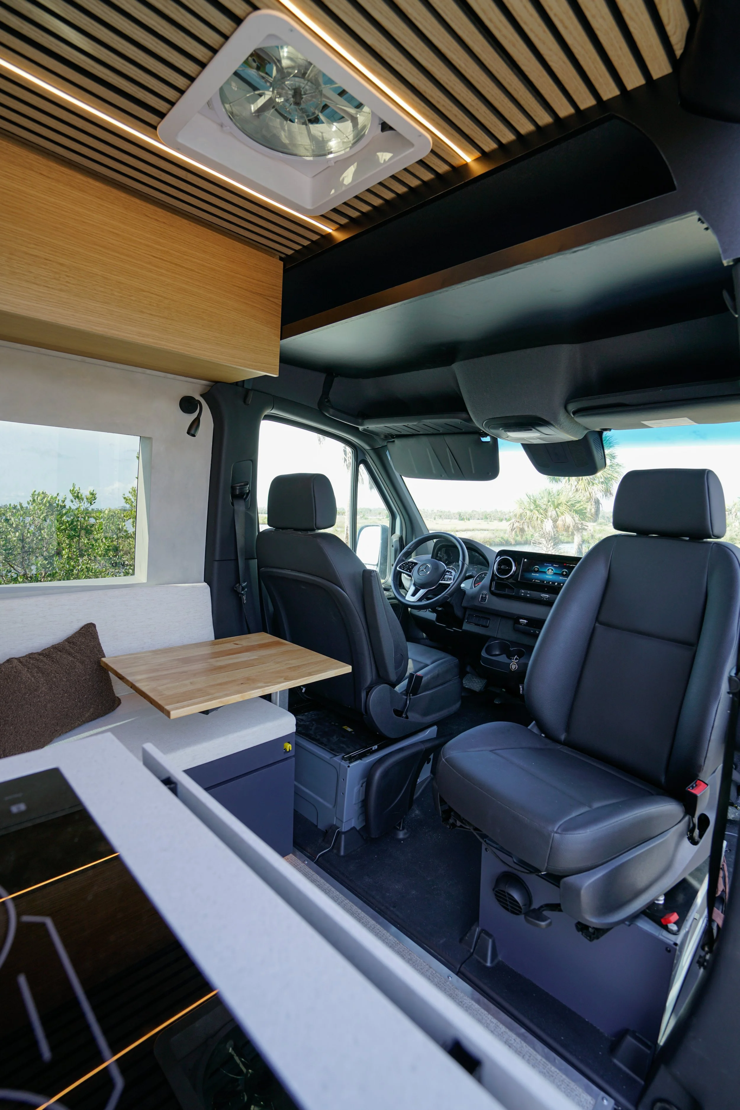 Interior of a camper van with a view of the driver's seat, dashboard, and a small seating area. Overhead, there are wooden and dark panels, a ceiling light, and a window showing an outdoor landscape with trees and a clear sky.