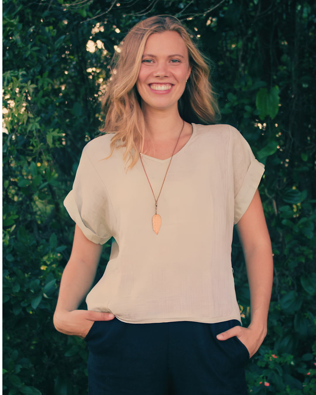 Smiling blonde woman wearing a beige blouse, pendant necklace, and dark pants outdoors against lush green foliage