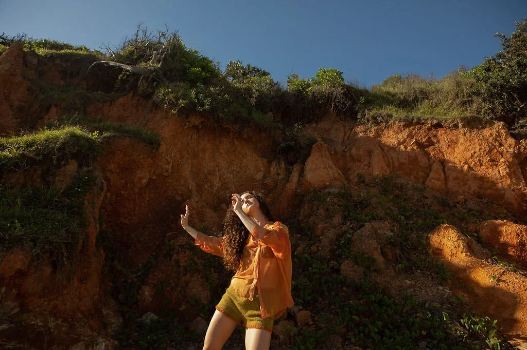 A woman with curly hair wearing an orange jacket and yellow shorts standing outdoors near a rocky cliff, with blue sky and green foliage in the background.