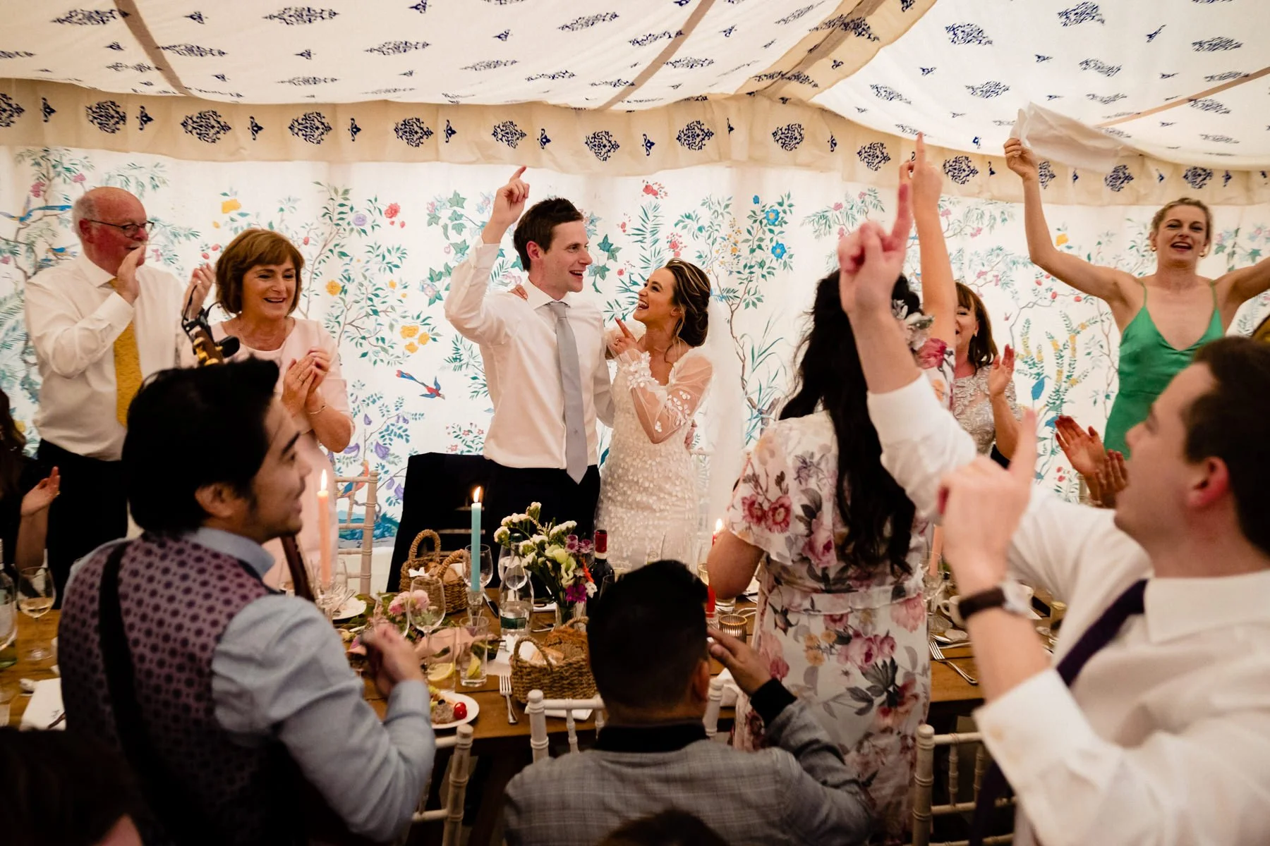 People celebrating at a wedding reception in a decorated tent, with a bride and groom at the center, surrounded by guests clapping and raising hands.