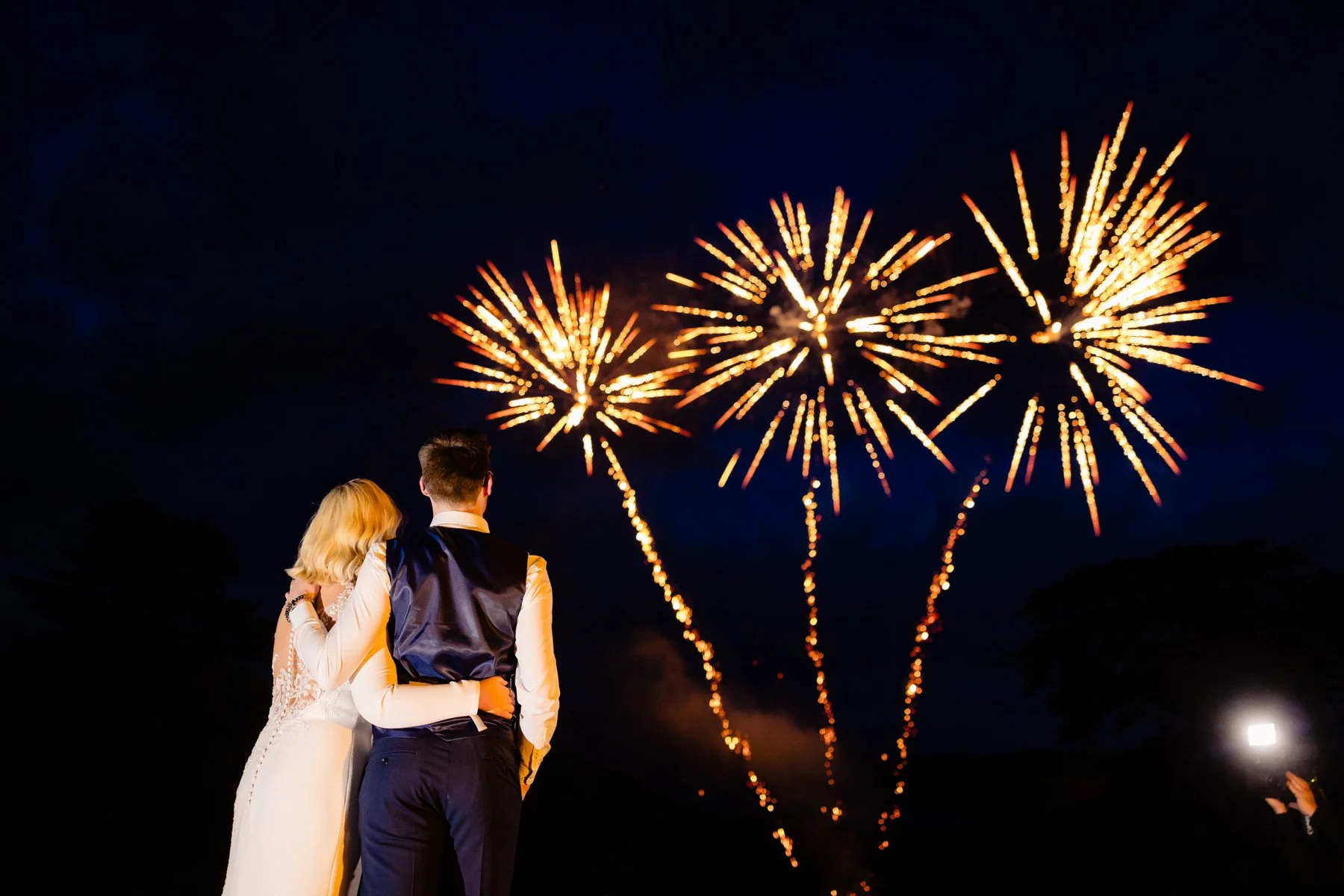 Couple watching fireworks at night, with the woman in a white dress and man in formal attire.