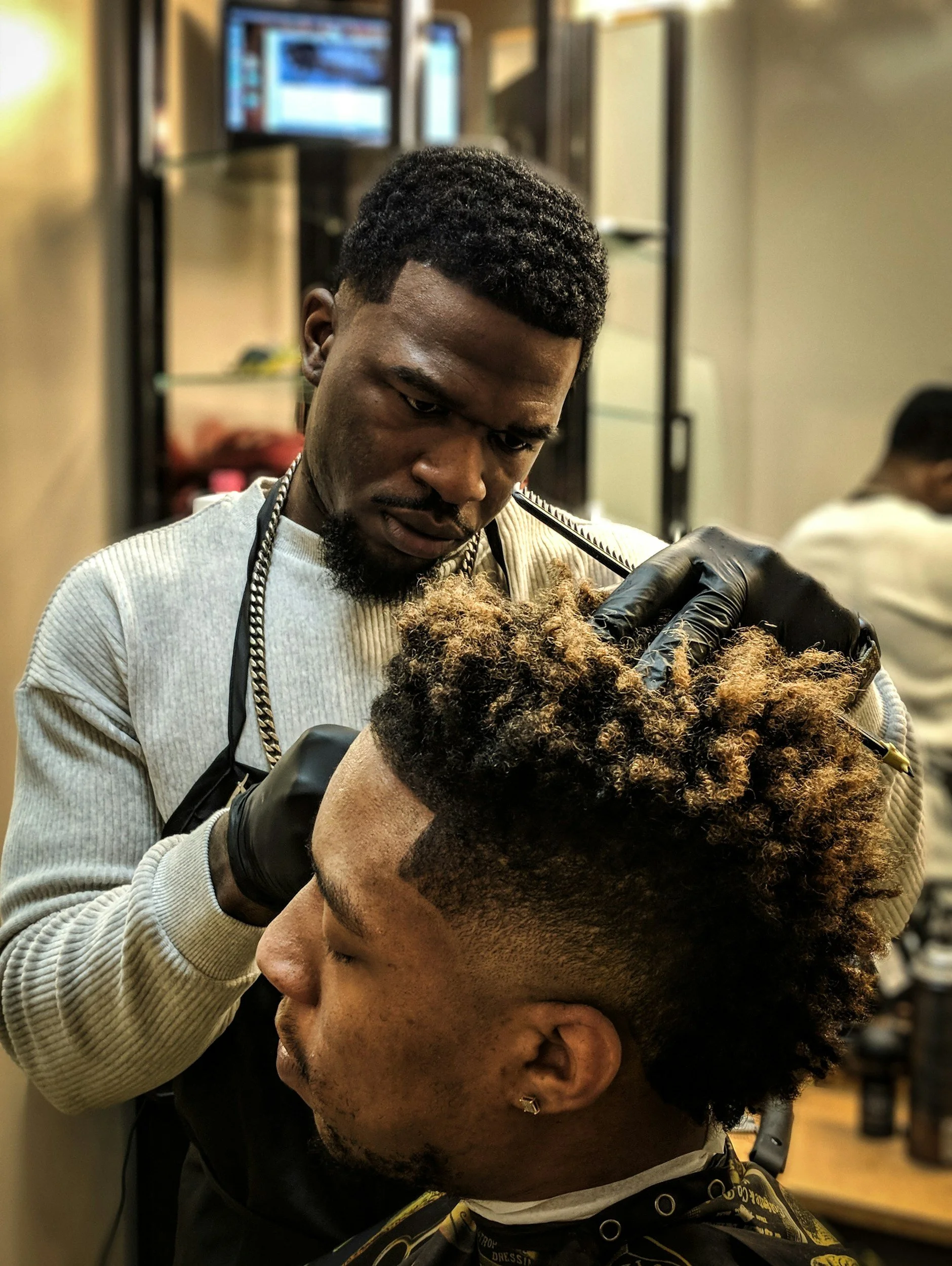 Barber in a gray sweater and black gloves carefully cuts a man's hair with a clipper, creating a fade hairstyle in a barbershop setting.