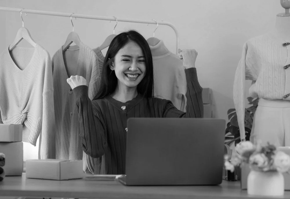 A woman smiling and celebrating with her arms raised in front of a laptop, in a clothing store or boutique with hanging sweaters and clothing mannequins.