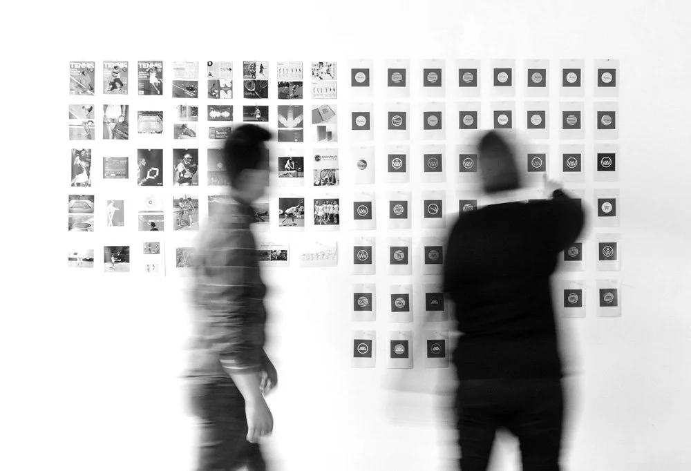 Two people look at a wall display of numerous small images and icons in an art gallery or exhibition space.
