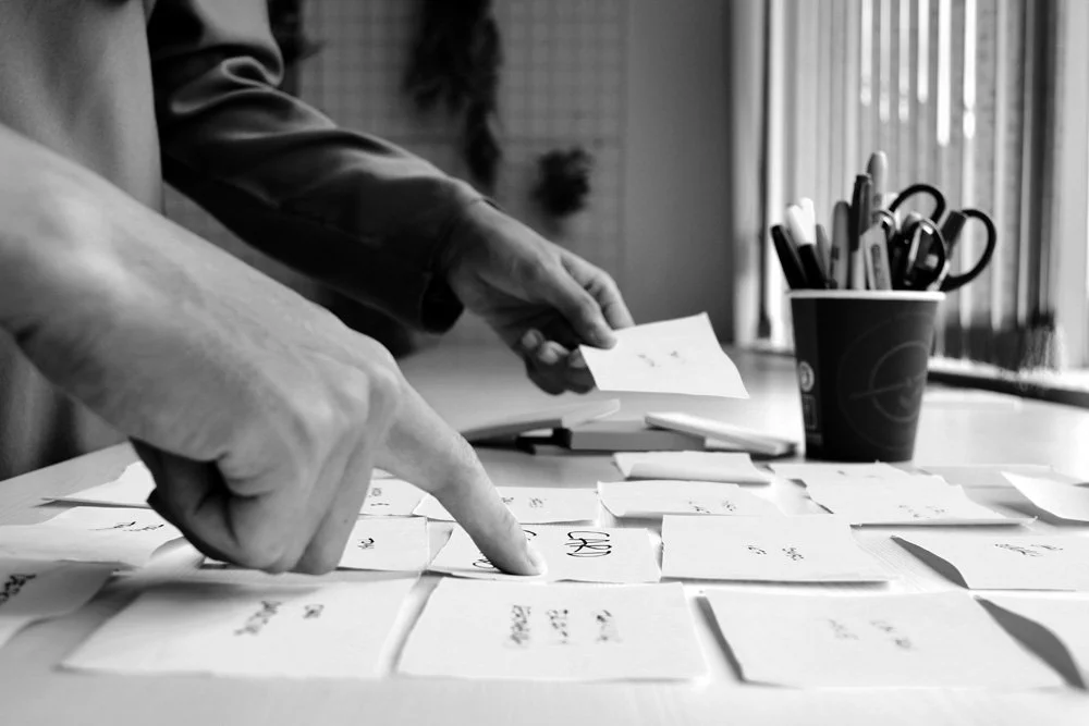 Hands sorting through various handwritten notes or cards on a table, with a container of writing utensils in the background.