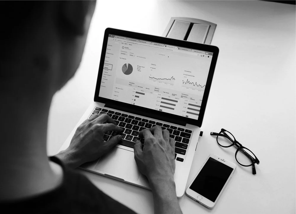A person using a laptop displaying analytics and graphs, with a smartphone and glasses on the desk.