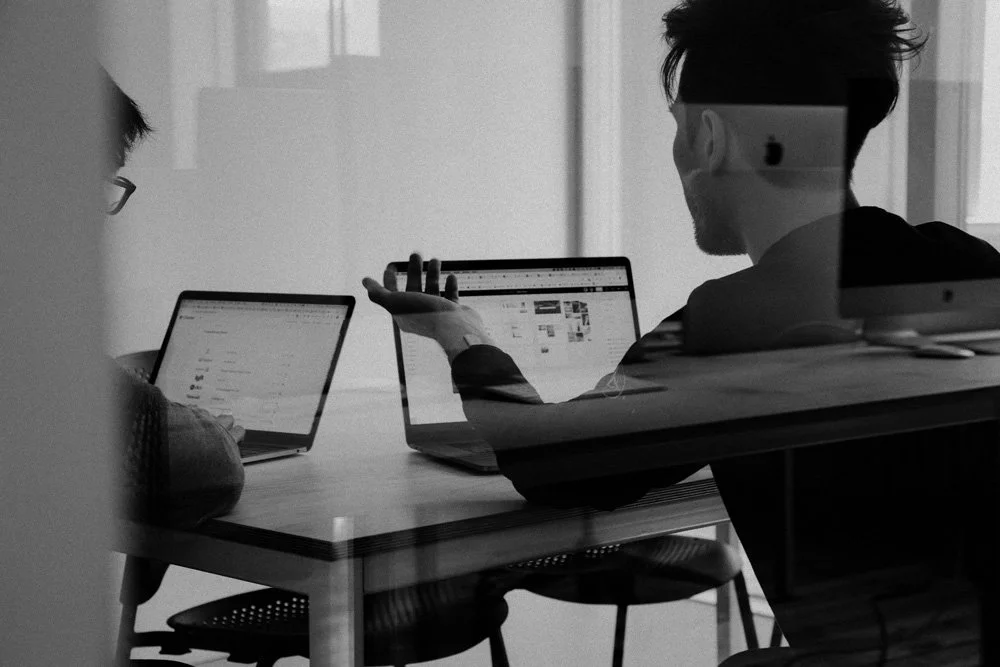 Two people sitting at a table working on laptops, viewed through a glass partition, in a black and white photo.