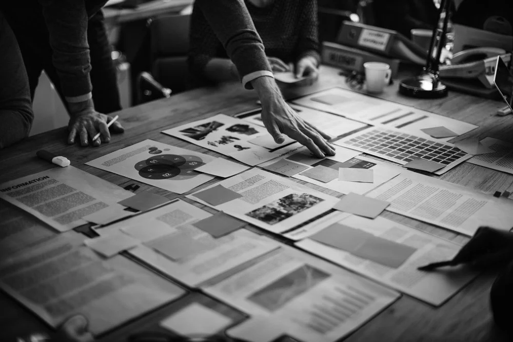 People discussing over a desk covered with documents, charts, and reports in an office setting.