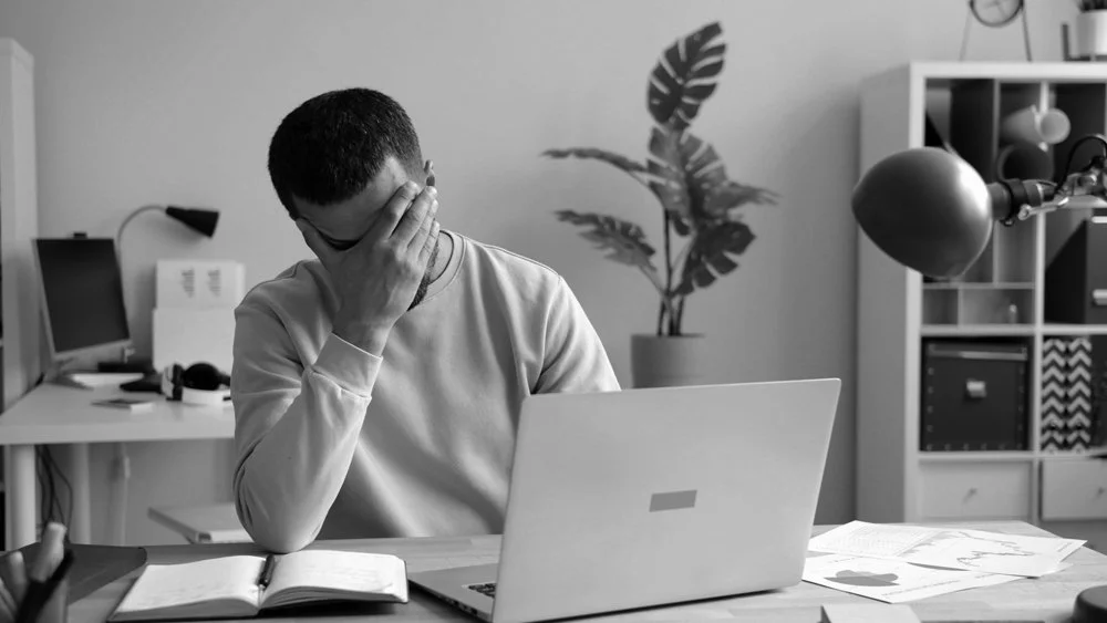 A person sitting at a desk with a laptop, holding their head in hand, appearing frustrated or stressed, with papers and an open notebook on the desk.