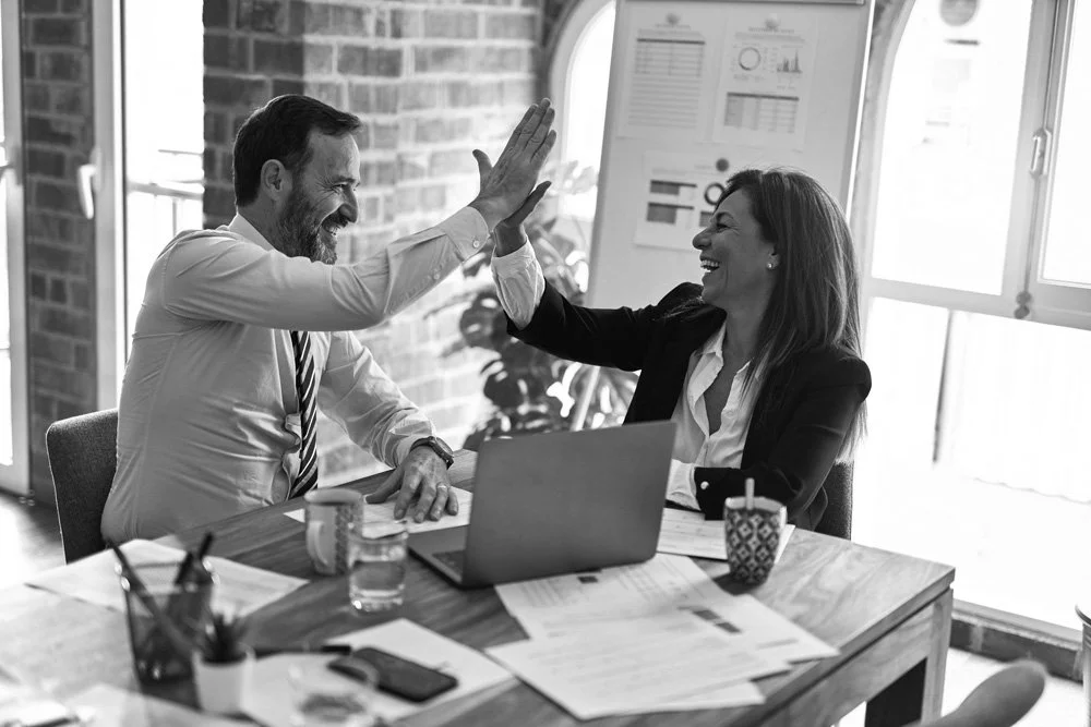Two business professionals, a man and a woman, giving each other a high-five in an office setting with documents, a laptop, and coffee cups on the table.