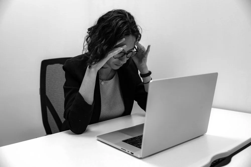Black and white photo of a woman with curly hair and glasses sitting at a desk, holding her head in frustration while looking at a laptop.