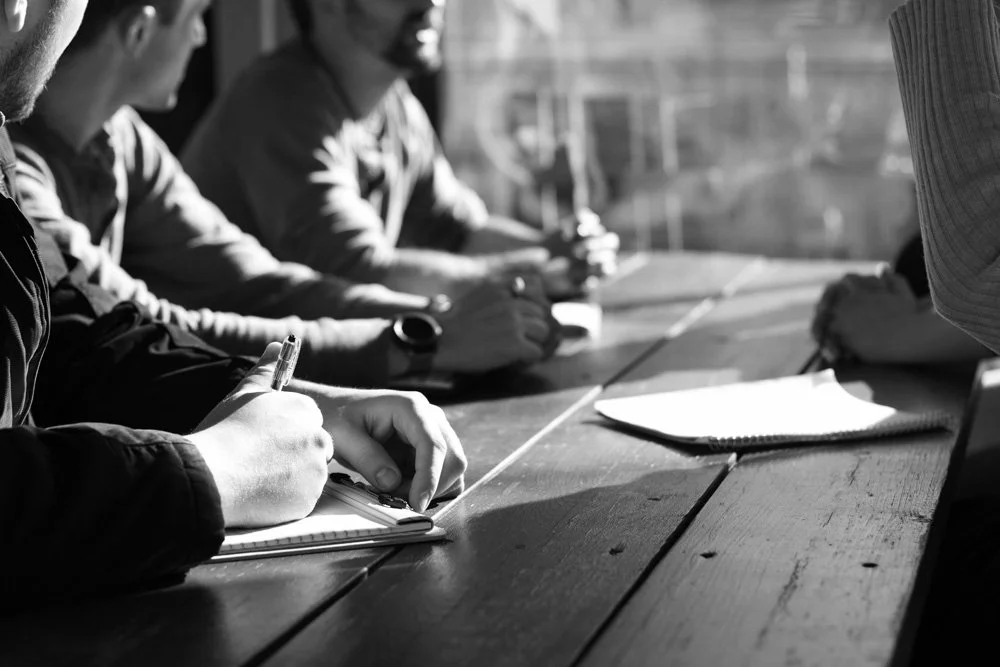 People sitting at a wooden table, taking notes in notebooks during a meeting or discussion.