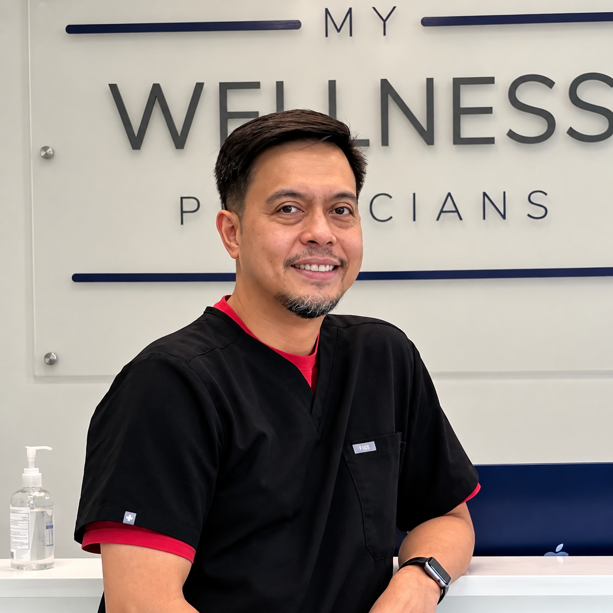 A man with short dark hair and a goatee, smiling and wearing a black shirt with red accents, standing in front of a sign that reads 'My Wellness Physicians', with a bottle of hand sanitizer on a white counter in the foreground.