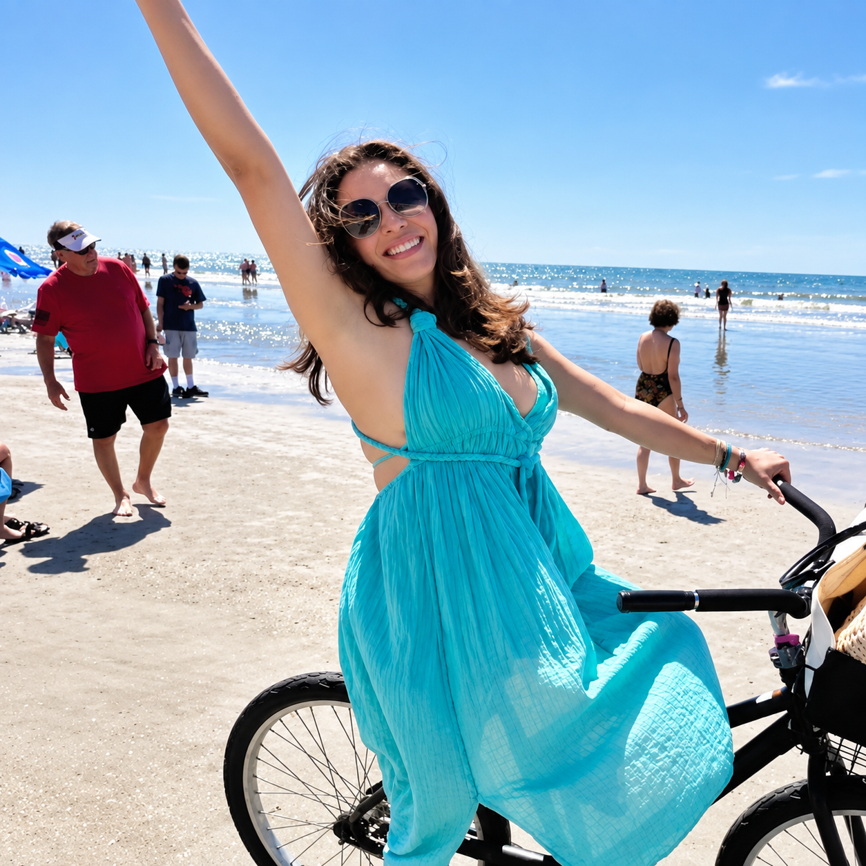 A woman in a blue dress riding a bicycle on a beach, smiling, with waves and other people in the background.