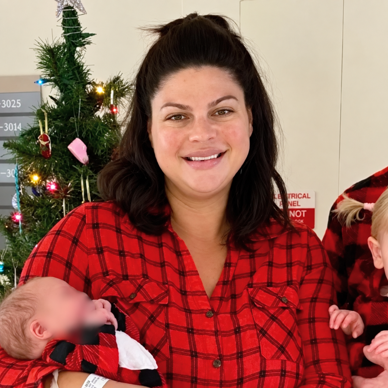 A woman with shoulder-length dark hair and light skin, smiling, wearing a red plaid shirt, standing in front of a decorated Christmas tree.
