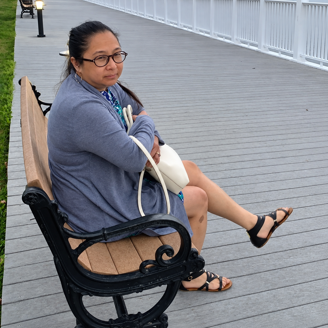 A woman wearing glasses, a blue jacket, and sandals sitting on a wooden bench by a boardwalk, holding a white bag, with her arms crossed and a sad facial expression.