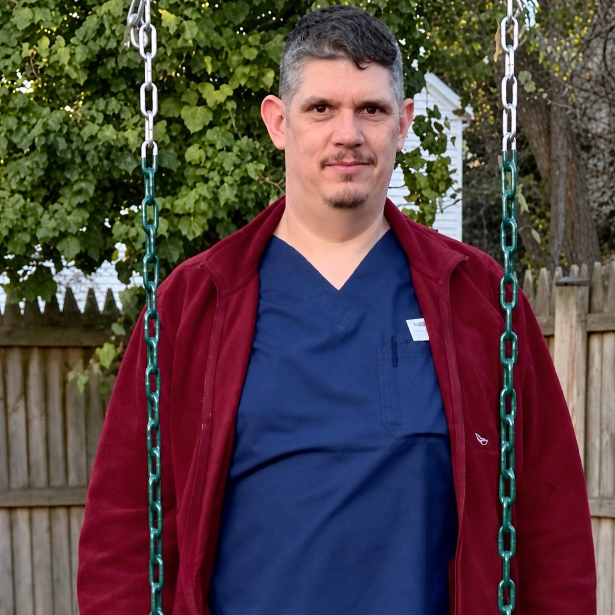 A man with short dark hair and a goatee standing outdoors in front of a wooden fence and trees, wearing a blue V-neck shirt and a red jacket, smiling at the camera.