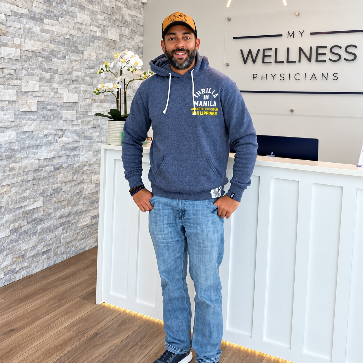 Man standing at a wellness clinic reception desk, wearing a blue hoodie, jeans, and a cap, smiling at the camera.