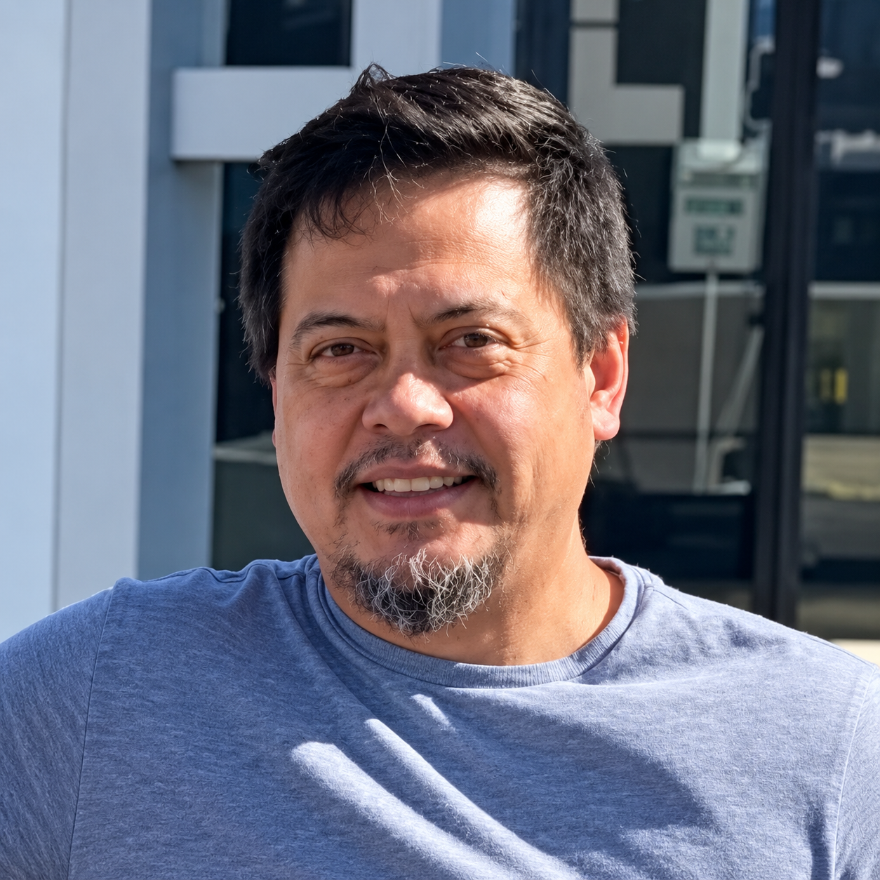 A smiling man with short dark hair and a goatee, wearing a gray shirt, outdoors with a building and electrical boxes in the background.