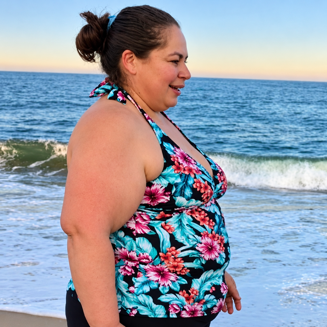 Woman with dark hair in a bun, wearing a floral swimsuit, standing on the beach with ocean waves in the background.