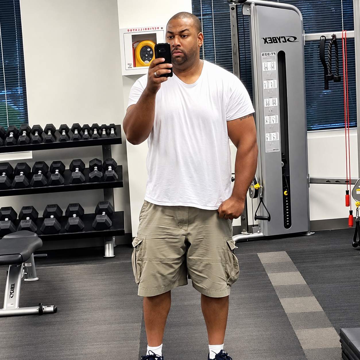 A man taking a mirror selfie in a gym, wearing a white t-shirt and beige shorts, with gym equipment in the background.