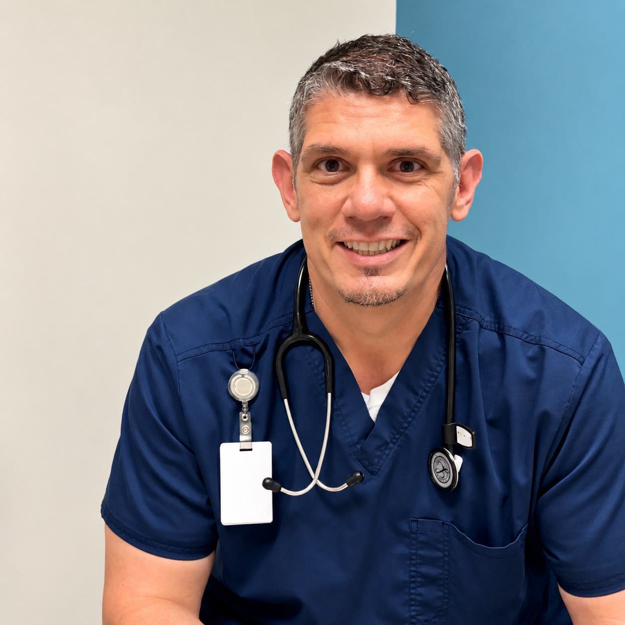 A smiling male nurse in navy scrubs with a stethoscope around his neck, posing in front of a beige and blue wall.