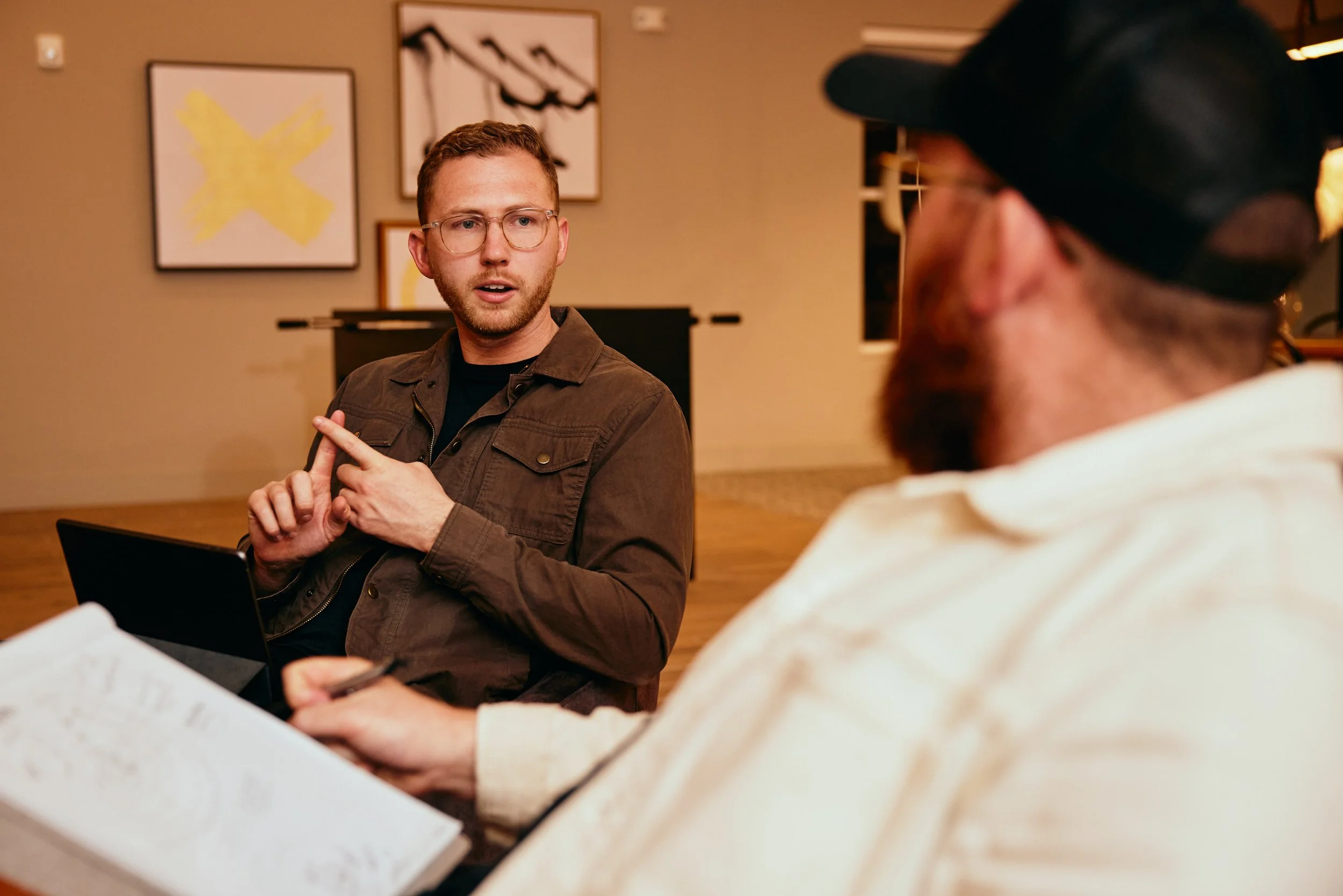 Two men having a conversation, one with glasses and a brown jacket, and the other with a beard and cap, sitting in a room with abstract art on the wall.