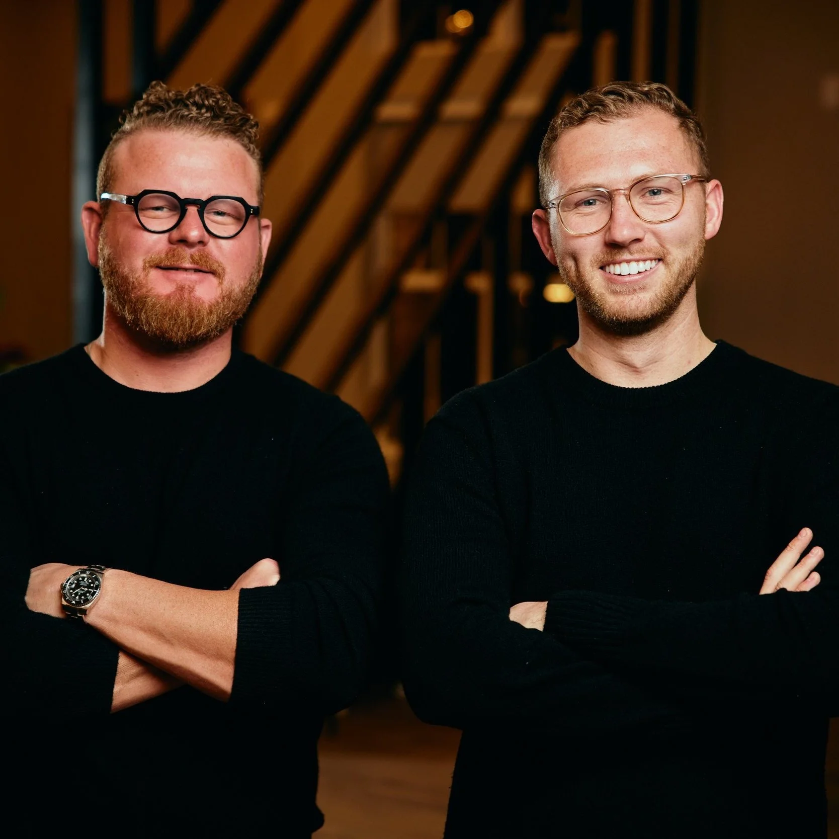 Two men with glasses and beards standing with arms crossed in a dimly lit modern interior space, smiling at the camera.