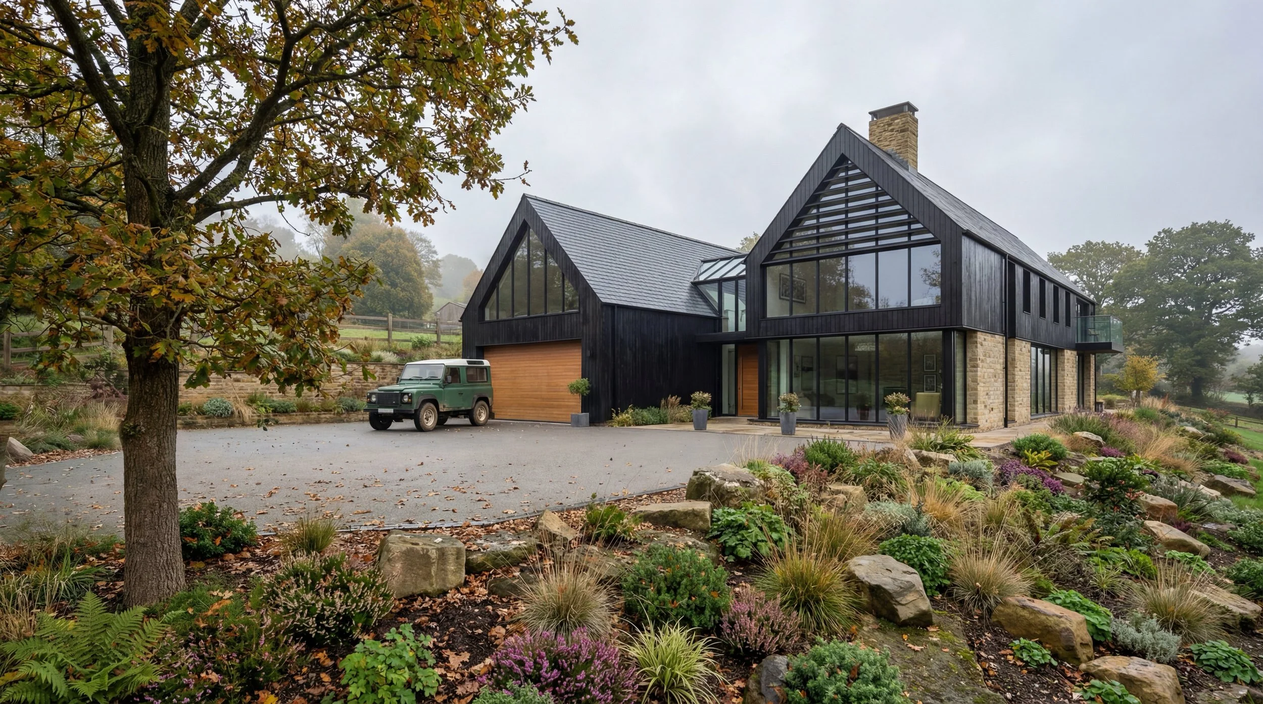 Modern black house with large glass windows, stone foundation, and wooden garage door amid a landscaped yard with trees, shrubs, and a vintage green Land Rover parked in the driveway.