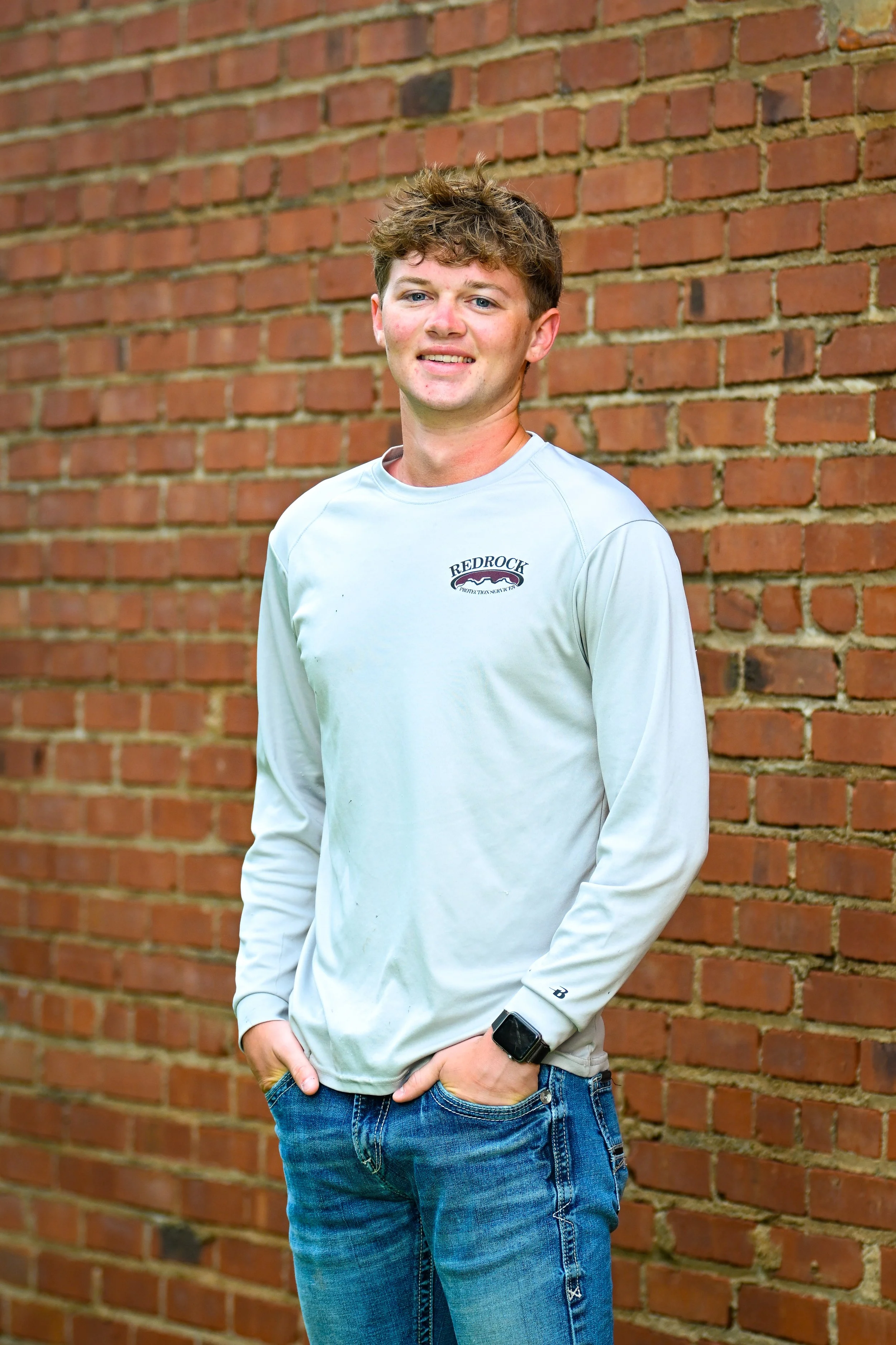 A young man with curly brown hair wearing a white long sleeve shirt and blue jeans, standing against a brick wall, looking at the camera with a slight smile.