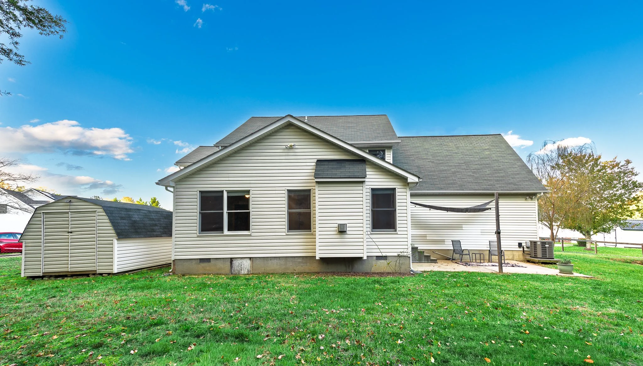 Backyard of a house with a lawn, a small storage shed, and outdoor seating area with a shade cloth, trees, and an air conditioning unit, under a bright blue sky with clouds.