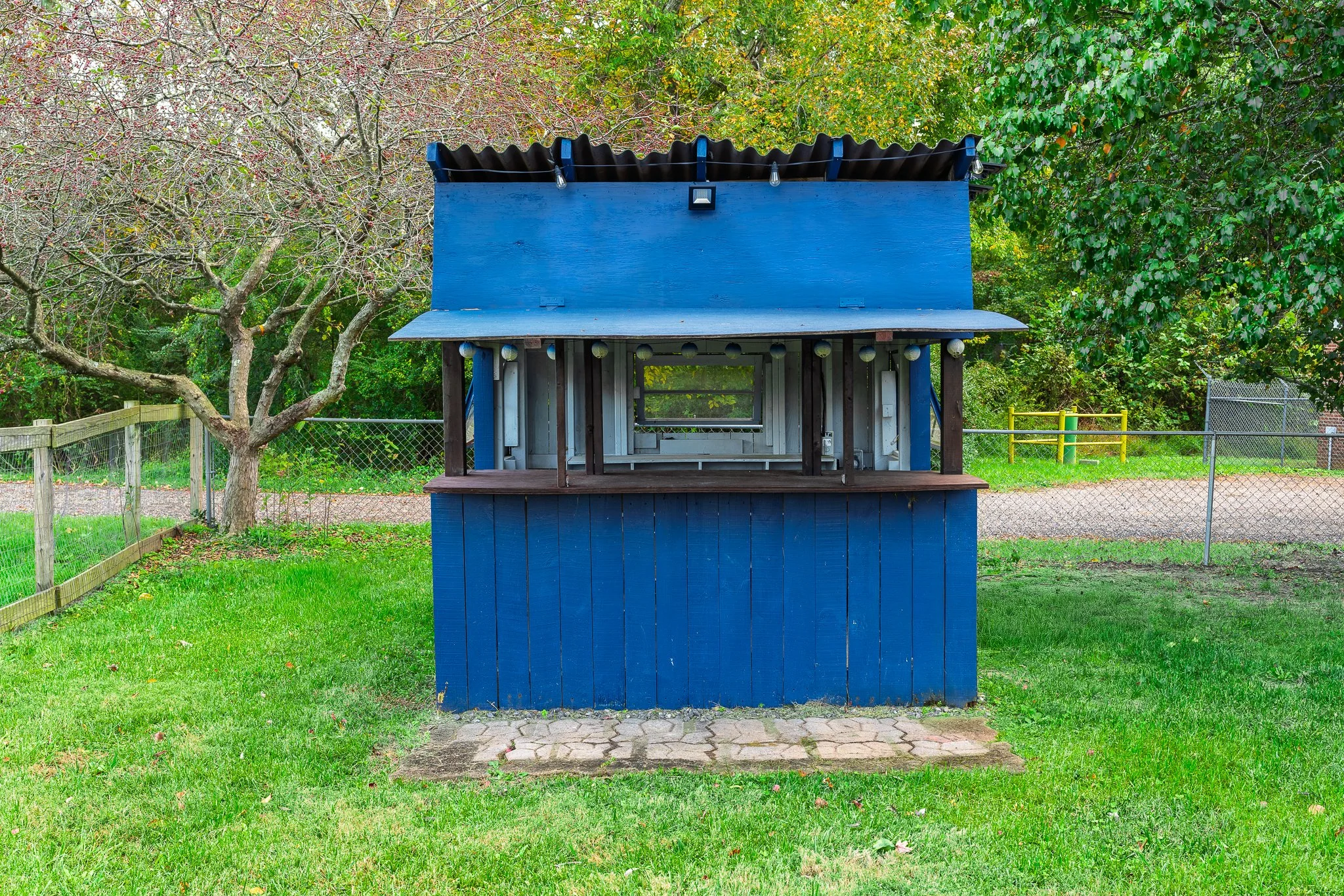 A small, blue, wooden playhouse with a porch, set in a grassy yard with trees and a chain-link fence in the background.
