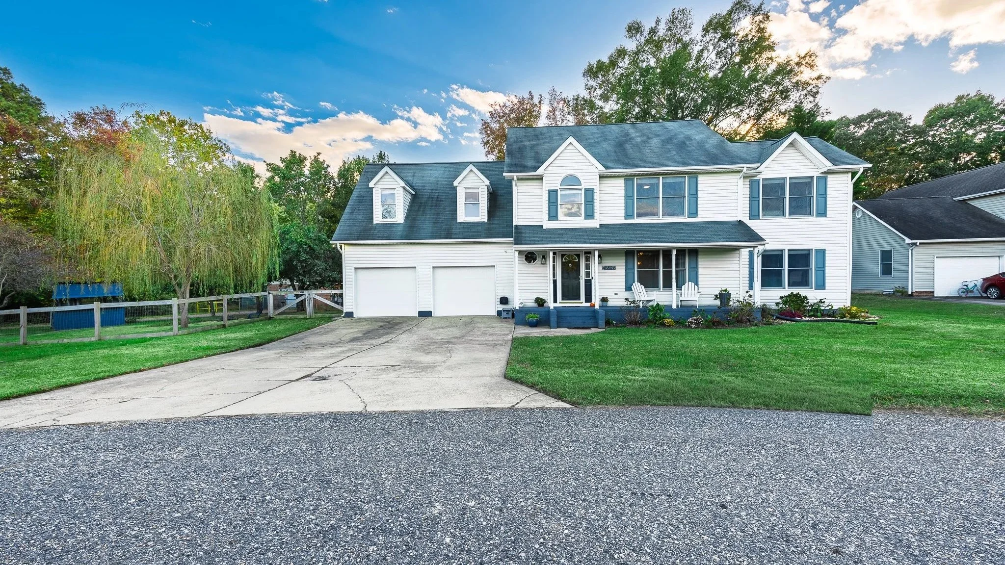 A two-story white house with blue shutters and a garage, front lawn with landscaping, and trees in the background under a partly cloudy sky.