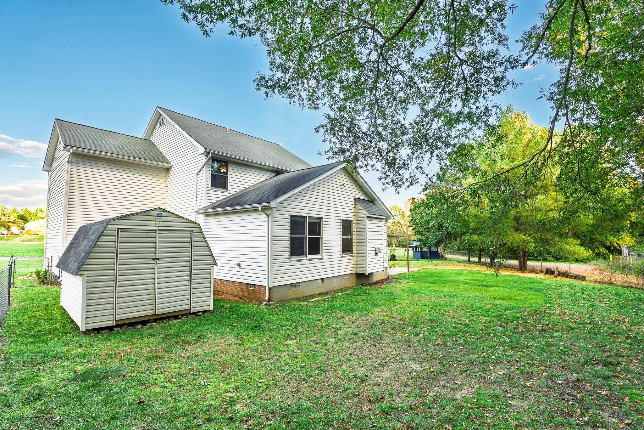 Backyard with white house, green grass, trees, and a storage shed under a blue sky.