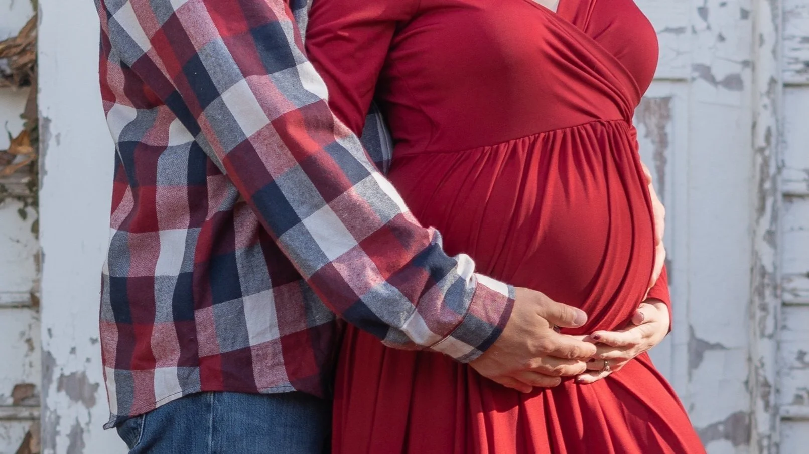 A pregnant woman in a red dress holding her belly with both hands, while a man in a plaid shirt gently holds her stomach from behind, standing outdoors in front of a weathered white wall.