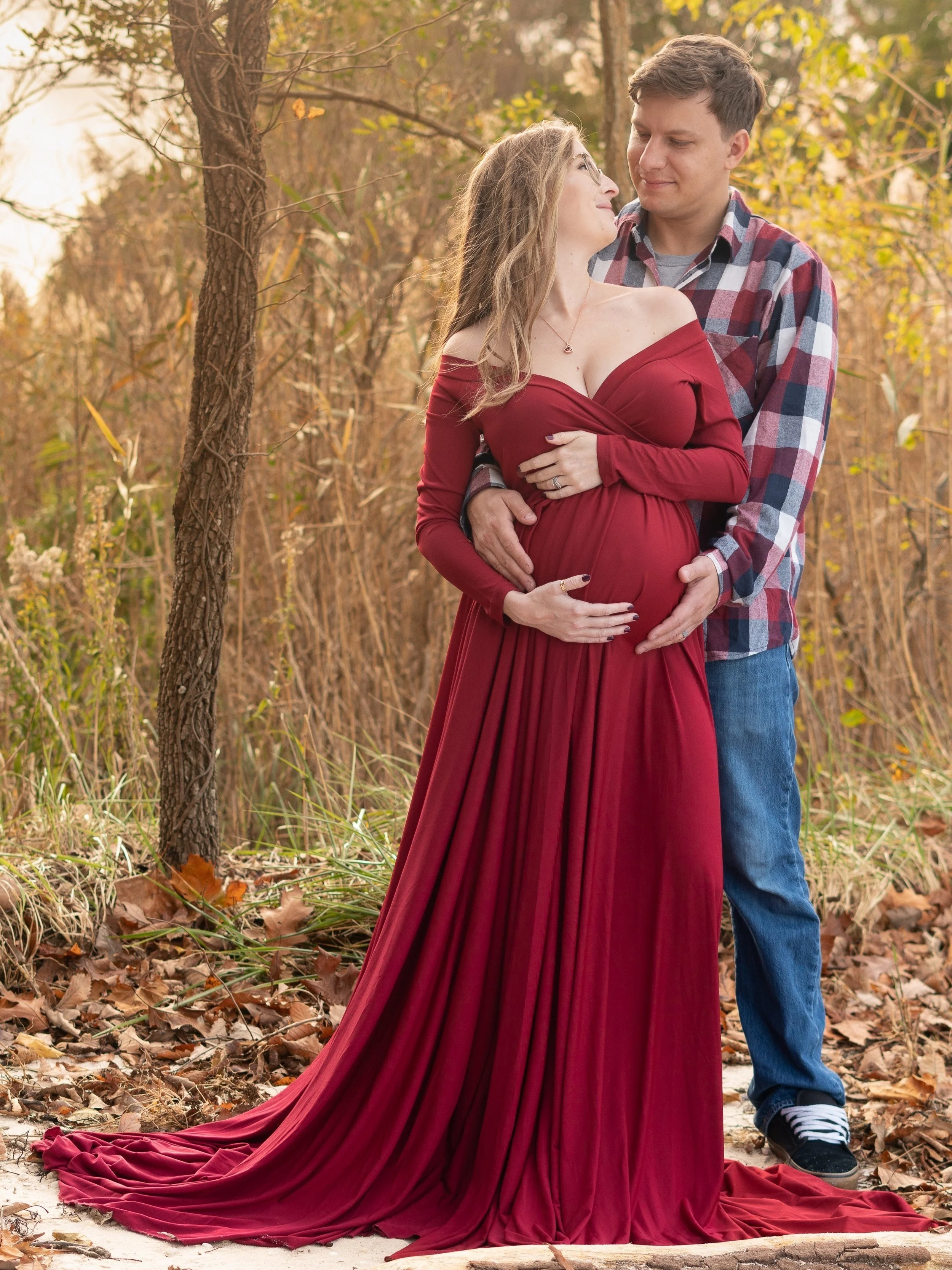 A pregnant woman in a flowing red dress stands outdoors with her partner, who is wearing a plaid shirt and jeans, both sharing an intimate moment in a natural setting with autumn foliage.