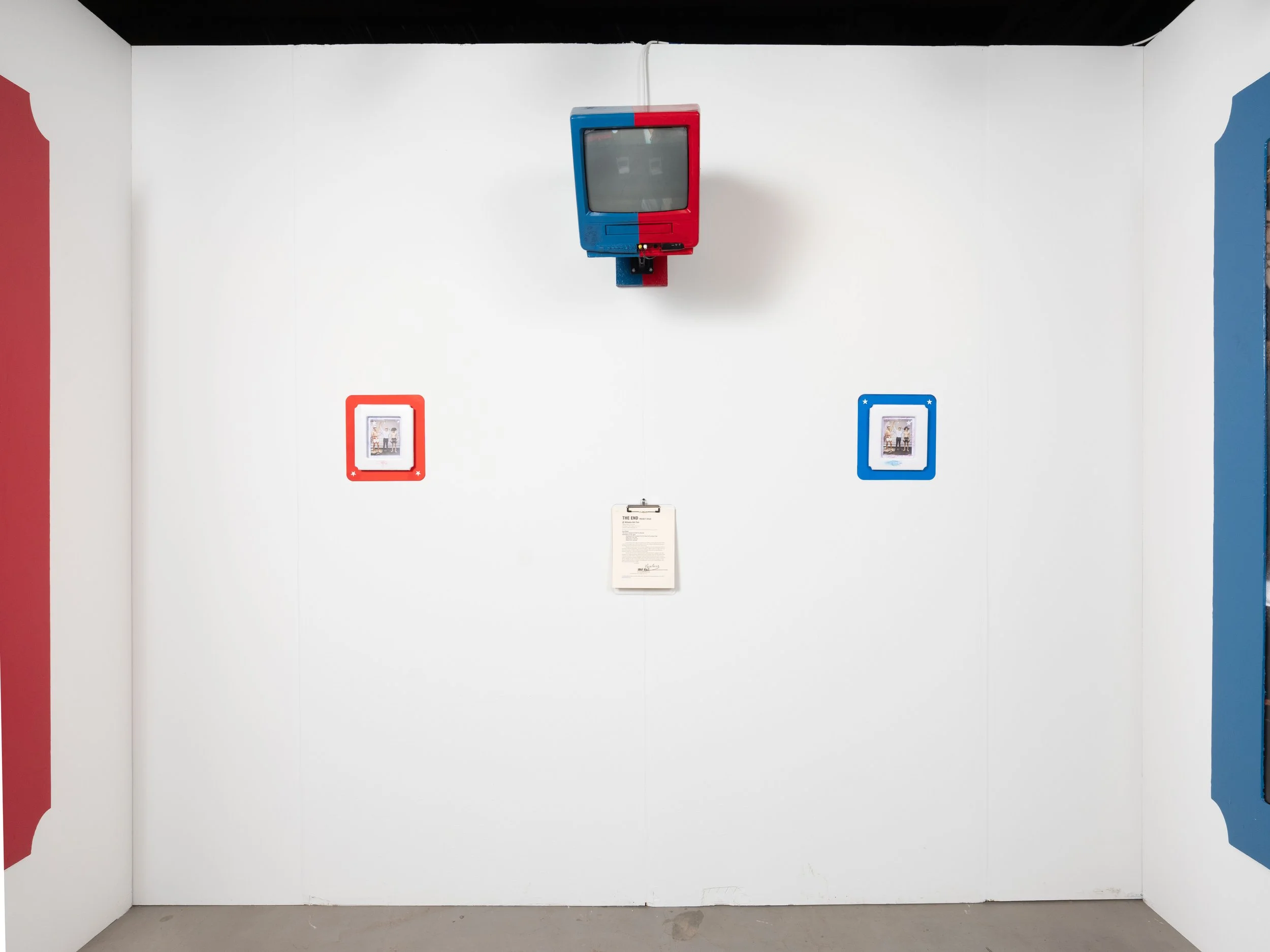 White gallery wall with a vintage television mounted at the top center, two small framed pictures below and to the sides, one in a red frame on the left and one in a blue frame on the right, and a clipboard hanging in the middle.