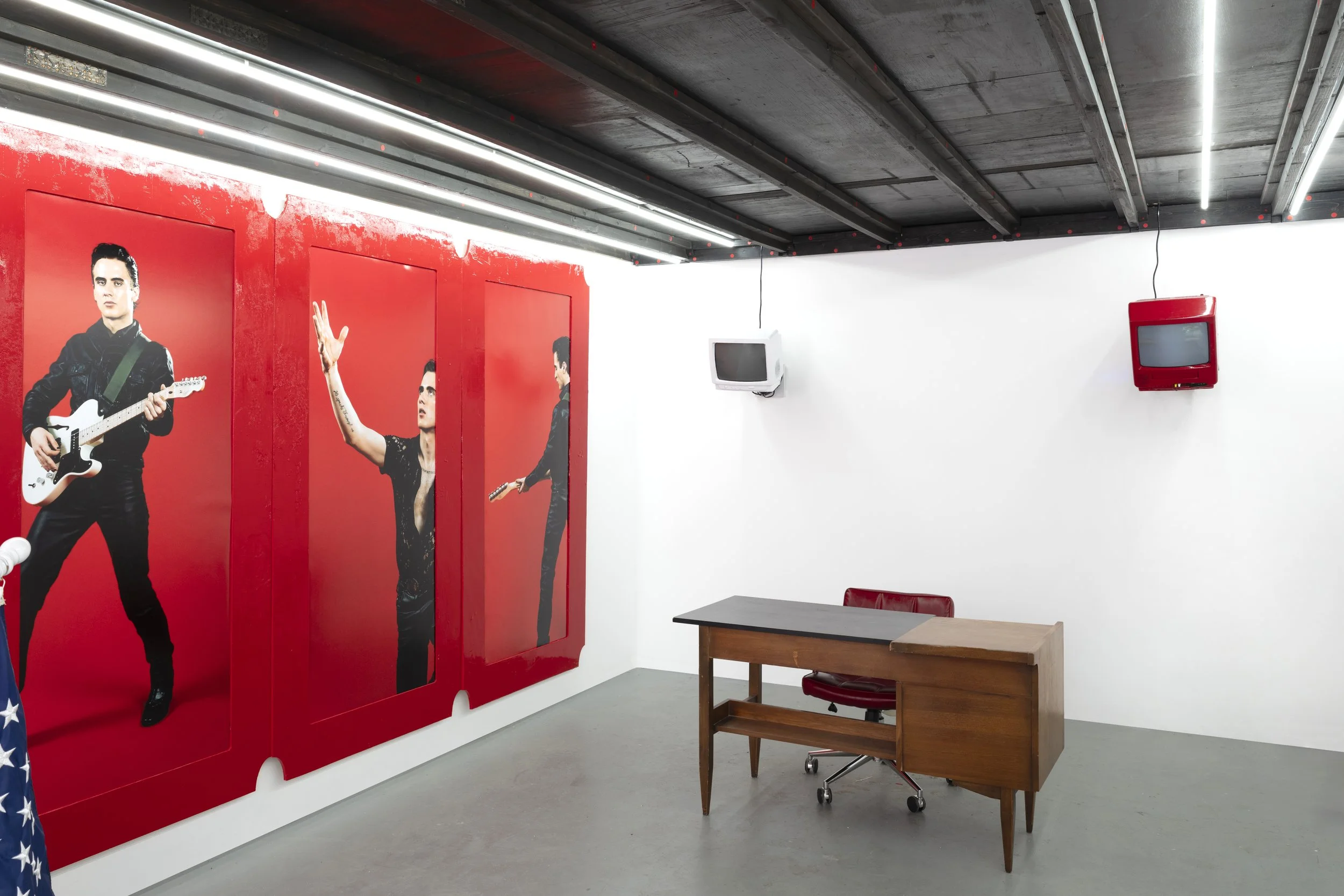 Interior of an art gallery with a large red mural of a man holding a guitar, a woman with tattoos and painted nails raising her hand, and another man playing a guitar in the mural. White walls, a small desk with a red chair, two small wall-mounted TVs, and a black ceiling with strip lighting.