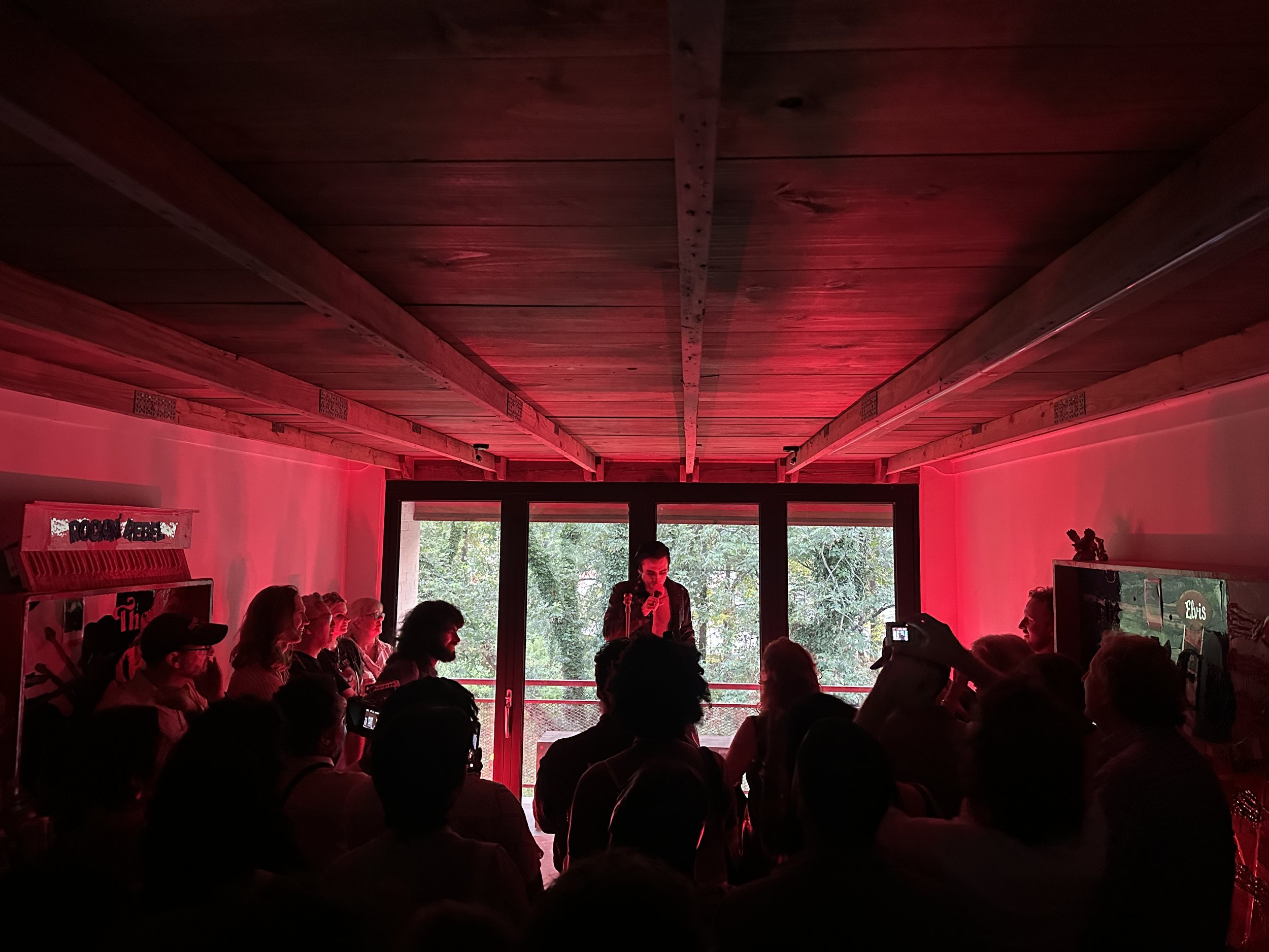 A singer performing on stage surrounded by an audience in a dimly lit room with red lighting, a wooden ceiling, and large windows showing trees outside.